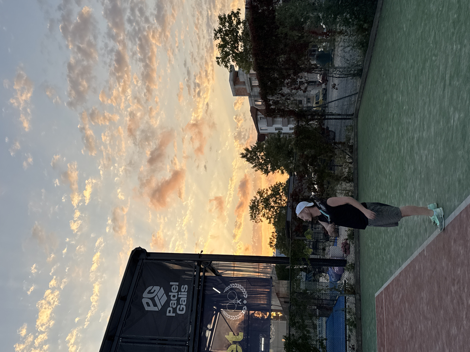 The image depicts a man standing on a padel tennis court at sunset in Tsarevo, Bulgaria. The sky is filled with dramatic clouds, illuminated by the setting sun, casting an orange and pink glow. The man, wearing a white cap, black tank top, grey shorts, and light blue sneakers, is holding a padel racket over his shoulder. He is looking upwards, towards the sky. To the left of the man, a large black structure with "Padel Galis" branding is visible. The court itself is divided into two sections: one reddish-brown and the other green. A fence surrounds the court, and beyond it, trees and buildings are visible. The overall scene captures a serene moment on the padel court as day transitions into evening in Tsarevo.