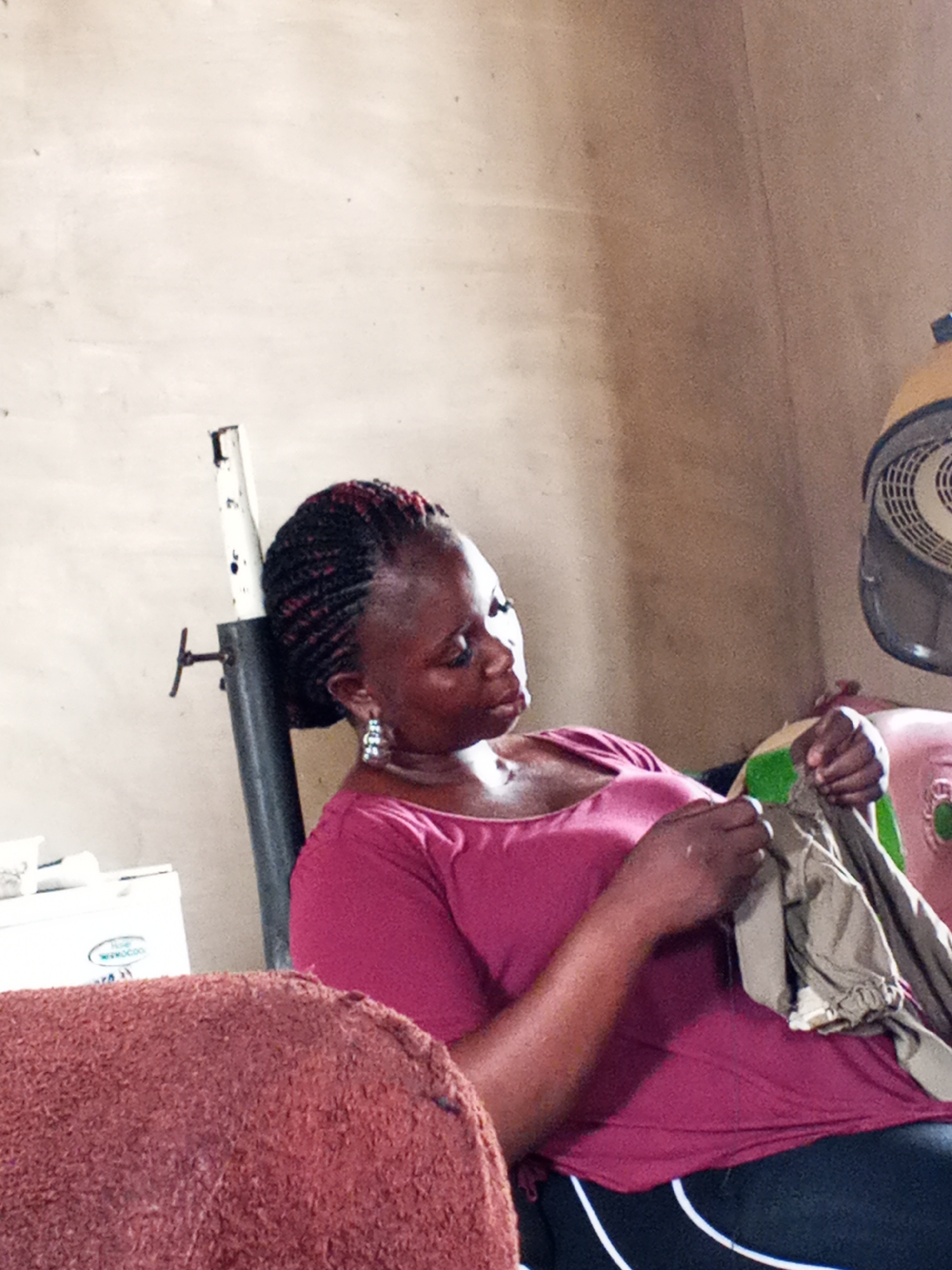 In an indoor space, a woman sits with braided hair, adorned with earrings. She wears a magenta top and dark pants with white stripes. The woman is focused on sewing a garment of neutral color, which she holds in her hands. To her left, there's a white object, possibly a cooler, and behind her, a metal support stand and a hair dryer. The setting suggests a residential area, possibly in the city of Adonkolo, Nigeria.