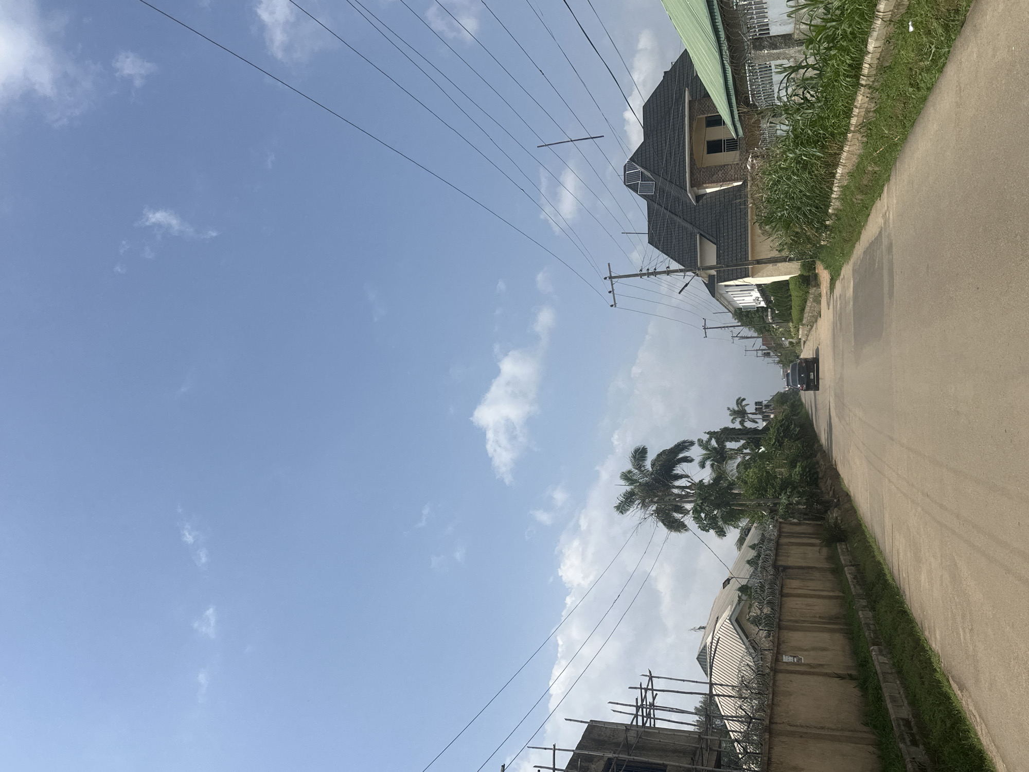 The image depicts a street scene in Abuja, Nigeria, under a bright, cloudy sky. The perspective is from a low angle, looking upwards and slightly to the right. A paved road occupies the right side of the frame, bordered by a strip of green grass and a concrete wall topped with razor wire.
Along the road, several buildings are visible. The most prominent is a multi-story house with a dark, sloped roof, featuring windows and what appear to be solar panels. Further down the street, more houses and a building under construction with scaffolding are present. Palm trees are interspersed with other lush greenery, indicating a tropical or subtropical climate.
Several power lines crisscross the sky, supported by utility poles, suggesting an urban or semi-urban environment with electricity infrastructure. A black car is parked or driving on the road, and another vehicle is visible further in the distance.
The time of day appears to be daytime, likely late morning or early afternoon, given the brightness of the sky and the shadows. The weather is partly cloudy, with patches of white clouds against a pale blue sky. There is no visible text in the image. The scene conveys a sense of everyday life in a Nigerian city.
