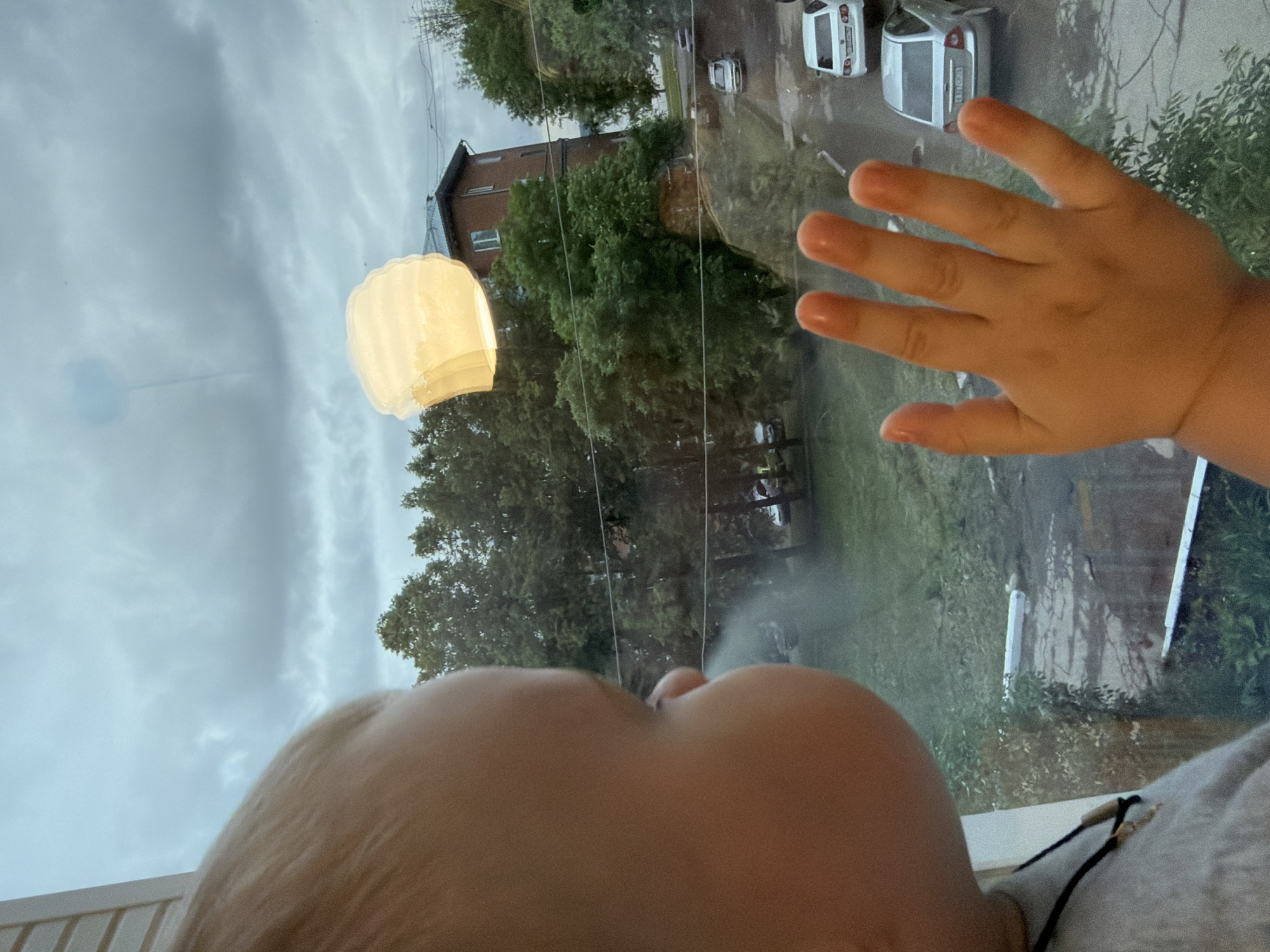 A low-angle, indoor photograph depicts a baby looking out a rain-streaked window. The baby's face, and hand pressed against the glass, are prominent in the foreground. Outside, the view includes trees, a multi-story brick building, and parked cars, suggesting a residential area in the city of Votkinsk, Russia. A white, lantern-like object is suspended in the sky. The sky is overcast.