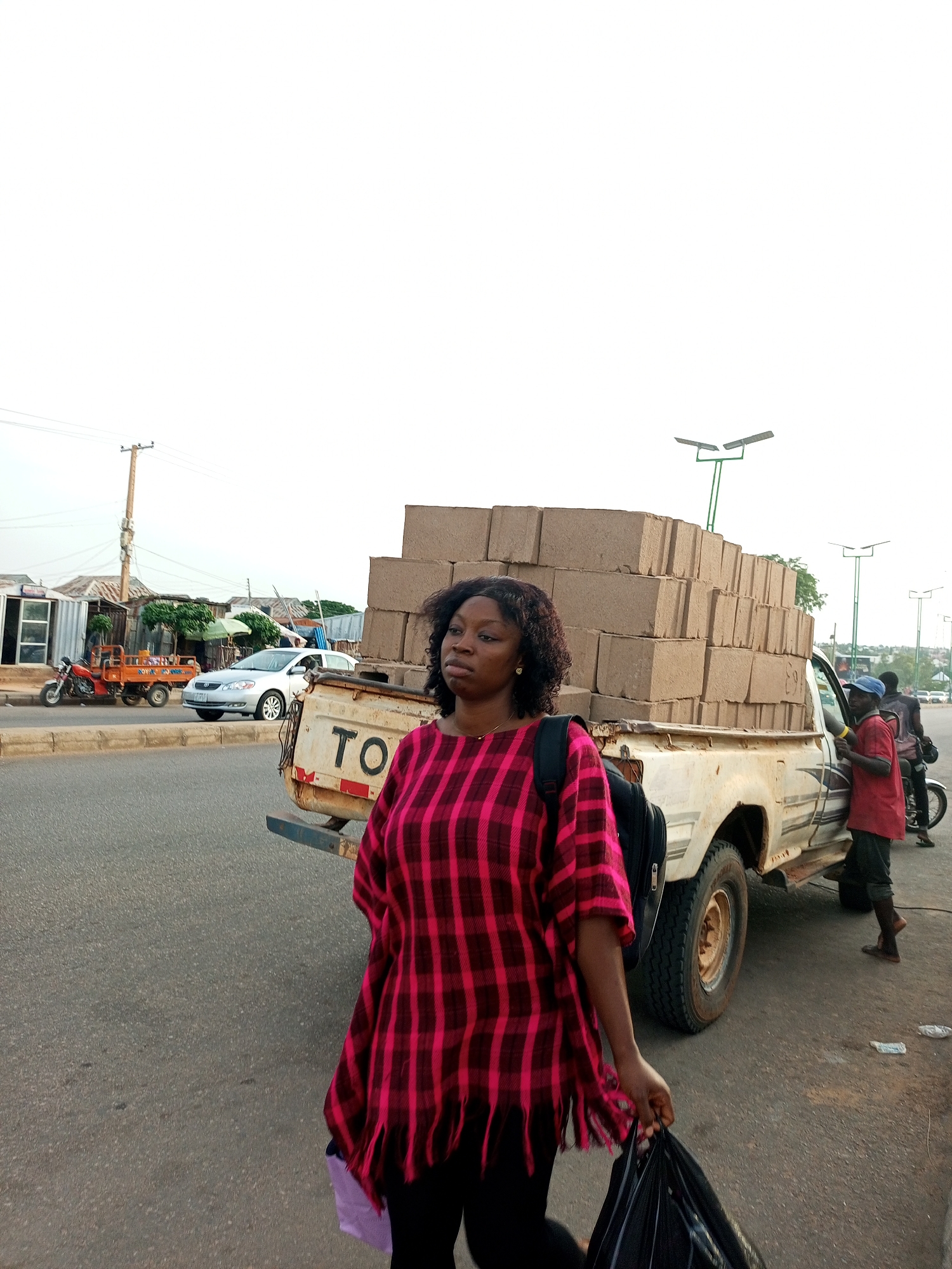 A medium shot captures a woman standing on a paved road in Tempure, Nigeria, looking towards the viewer with a neutral expression. She is wearing a red and black checkered, fringed tunic over black leggings and carrying a black plastic bag in her right hand. A black backpack is slung over her left shoulder. To her right, a white pickup truck is parked, its bed loaded with stacks of beige bricks. A man in a red shirt and blue cap stands near the truck, holding something yellow in his hand. Another person is partially visible behind him, wearing a dark shirt and dark pants, with a motorcycle to their left. The background shows a row of buildings with corrugated metal roofs and a utility pole. In the distance, more structures and trees are visible, along with streetlights. The sky is overcast.