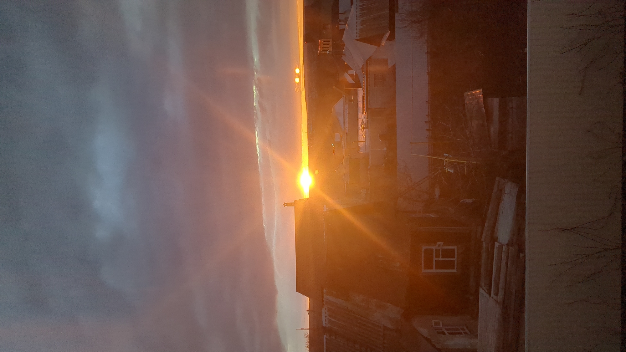 The image shows a view of a cityscape at sunset, possibly in Ulyanovsk, Russia. The sky is a blend of dark, stormy clouds and bright orange hues from the setting sun. The sun's glare dominates the horizon, casting long rays across the scene. The foreground is filled with rooftops and buildings, which appear silhouetted against the bright sky. The buildings are a mix of older structures with visible windows and newer constructions. Bare tree branches are seen in the right side of the frame. The image is taken from an elevated viewpoint.