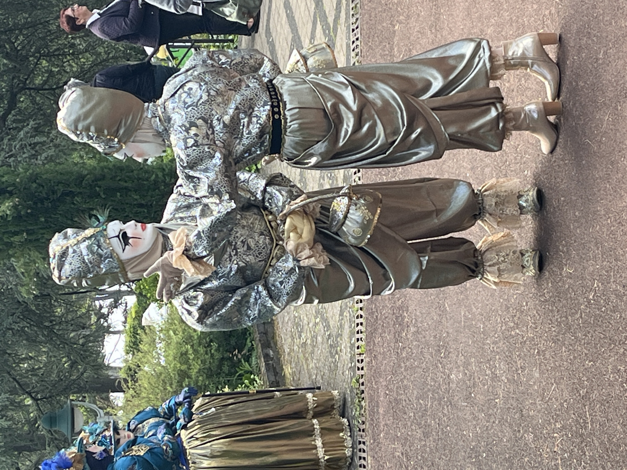 The image shows two people dressed in elaborate, old-fashioned costumes and makeup, characteristic of Venetian carnival figures. They stand close together on a paved surface, with greenery and other people in the background. The costumes feature silver and grey fabrics, lace details, and heeled boots. One figure holds a small purse. The scene appears to be outdoors, possibly a public space with trees and other decorative figures in the distance. Based on the context provided, it is plausible that the location is Saint-Galmier, France.