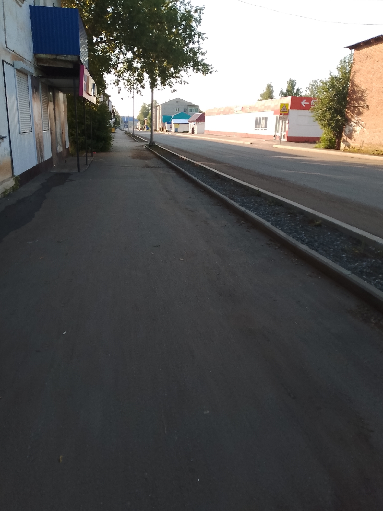 The image shows a street scene in Kranokholmskiy, Russia. On the left, a building with white siding and closed shutters lines the sidewalk. A blue awning extends over part of the building. A tree with dense foliage stands in front of the building, casting shadows on the sidewalk. The sidewalk leads to a street with a grey asphalt surface. To the right of the street, a long, narrow trench filled with gravel runs parallel to the road. Further down the street, several buildings are visible. One building is white with a red trim and features a sign with an arrow pointing to the left and a yellow triangular warning symbol. Another building to the far right is constructed of red brick. The sky is clear and bright. The scene appears to be a quiet urban area during daylight hours.