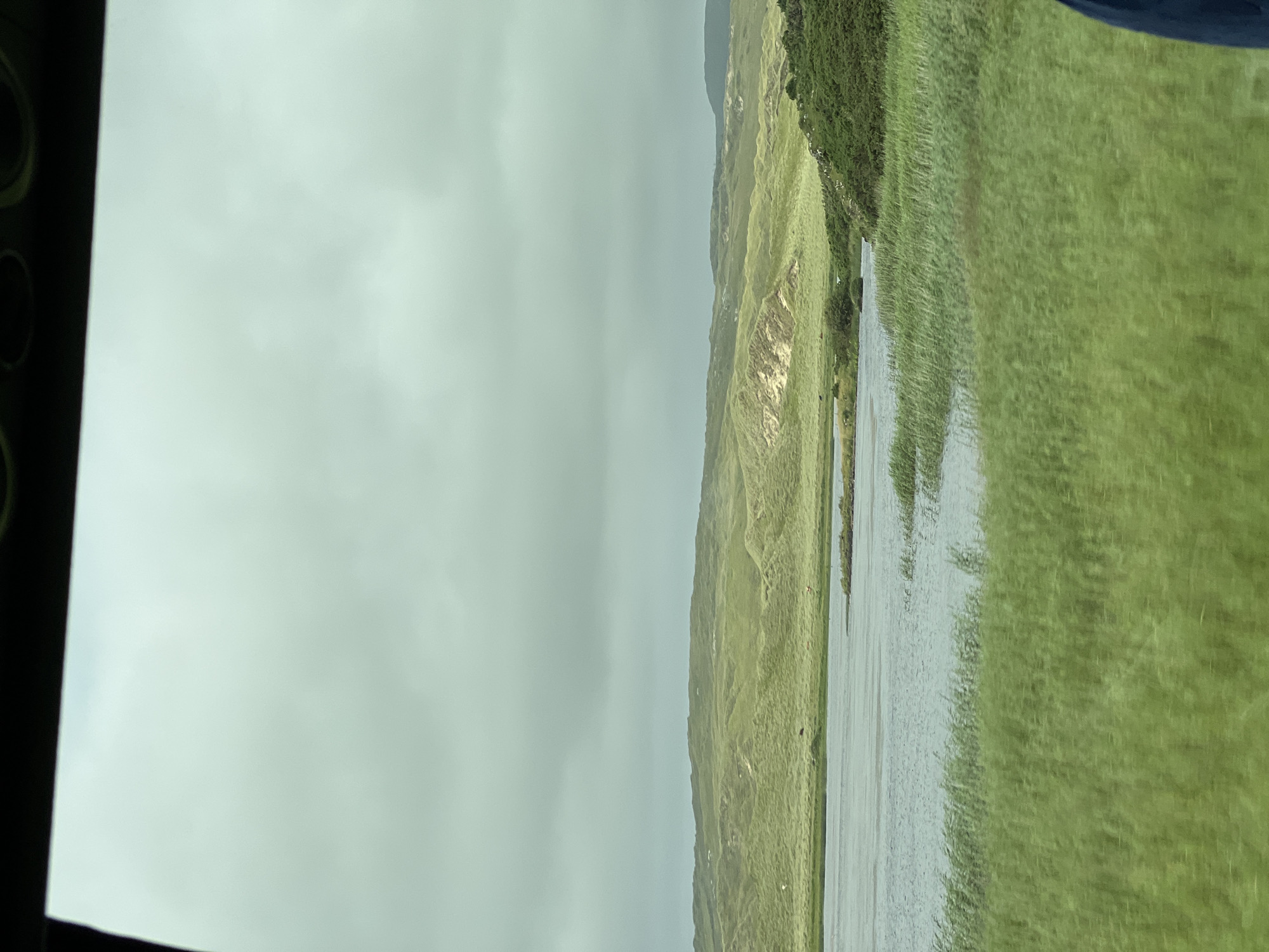 A scenic view of the coastline near Dunfanaghy, Ireland, showcases rolling green hills covered in grass and heather. The sky is overcast with gray clouds, suggesting a cool, potentially damp day. In the foreground, tall green reeds line the edge of a body of water, which appears to be a tranquil lake or inlet. The water reflects the muted sky, creating a serene atmosphere. Further back, the undulating landscape rises, dotted with sparse vegetation and sandy patches. The overall impression is one of natural beauty and a peaceful, remote location.