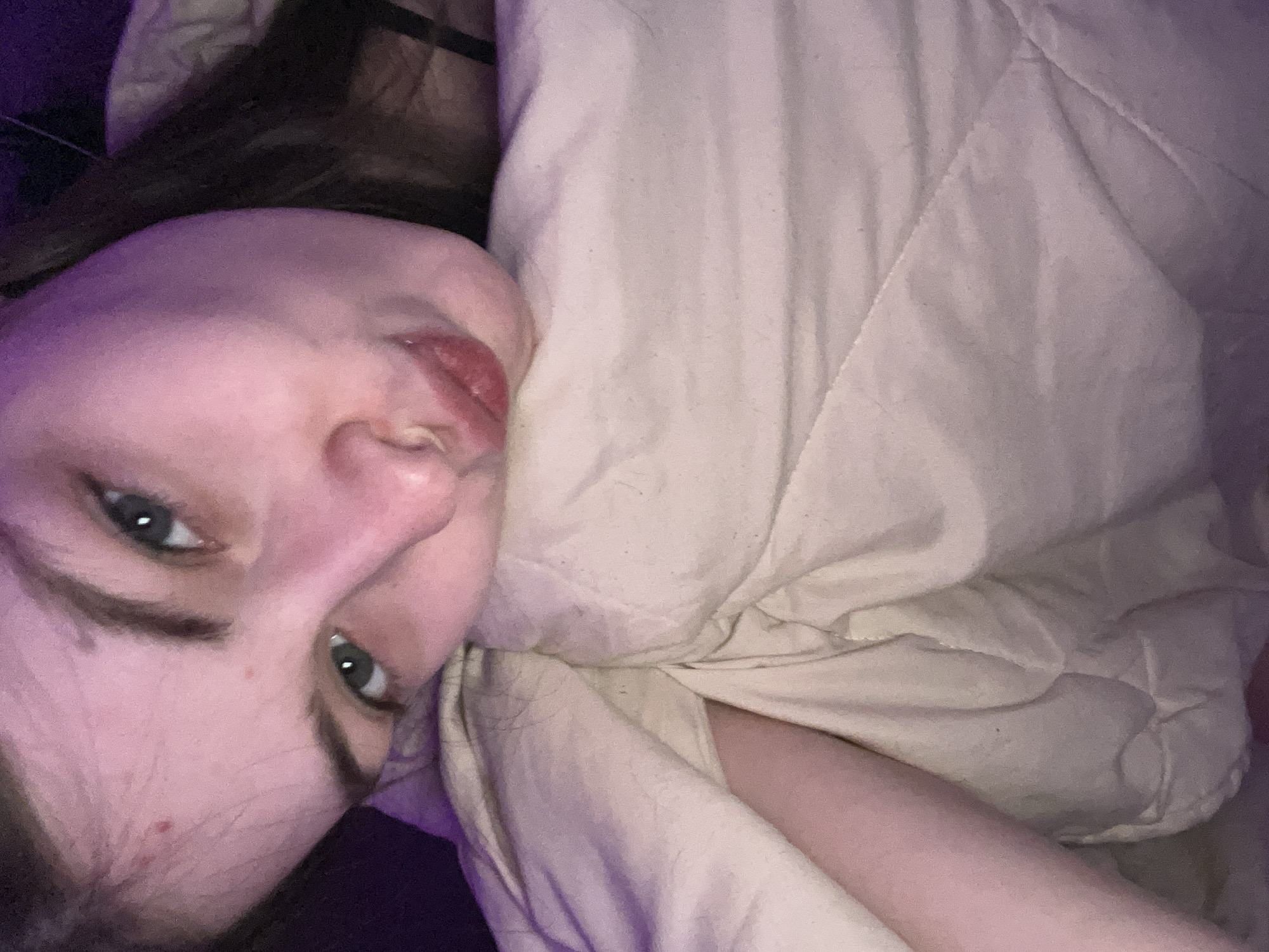 A young woman is lying in bed under a beige comforter. She is turned on her side, facing the camera. Her hair is dark. She has fair skin and gray eyes. The image is angled and has purple lighting. The location is not discernible from the image and cannot be determined to be Saint Petersburg, Russia.