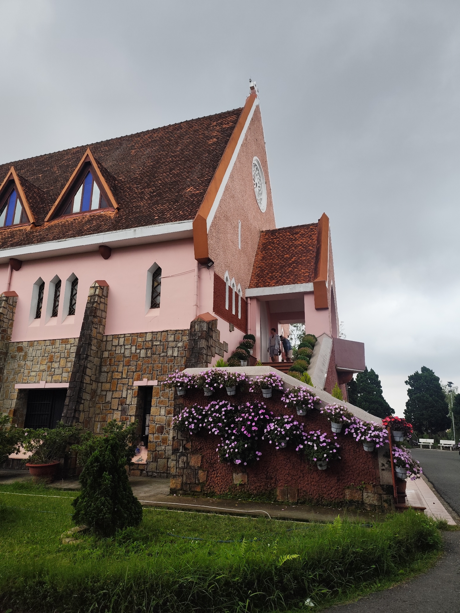 The image shows the exterior of the Cam Ly Cathedral in Da Lat, Vietnam, on an overcast day. The cathedral is painted a light pink and features a terracotta tile roof. The facade is adorned with stone accents and numerous triangular windows. A prominent feature is a wall covered in hanging planters filled with small pink flowers, cascading down from the steps of the entrance. Two individuals are visible near the entrance, one standing and looking down, the other bent over. The foreground consists of a grassy area with a small evergreen tree. To the right, a paved road is visible, with benches and trees in the background. The architecture appears to be a blend of Gothic and French colonial styles.