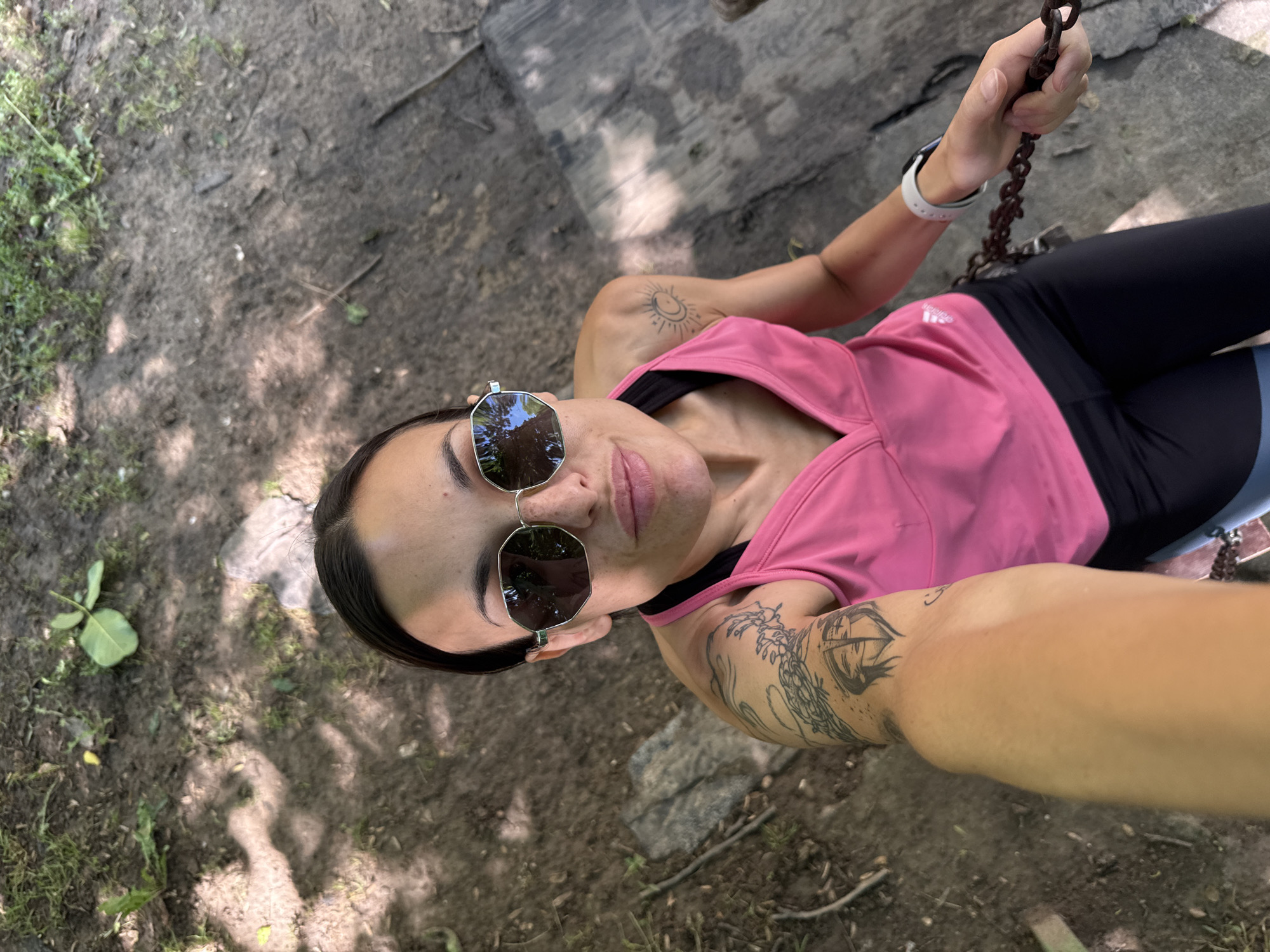 The image, taken in Pleven, Bulgaria, captures a woman from a high-angle perspective, looking directly into the camera as if taking a selfie. She is outdoors, likely in a natural setting, with a ground composed of dirt, sparse grass, and fallen leaves, illuminated by sunlight casting dappled shadows.
The woman has dark hair and is wearing silver-rimmed, octagonal sunglasses, reflecting the surrounding greenery. She is dressed in a vibrant pink sleeveless athletic top and black leggings. A white sports watch adorns her left wrist, and her right hand grips a rusty metal chain, suggesting she may be seated on a swing or similar structure.
Notable details include tattoos on both arms: a stylized sun and crescent moon design on her left shoulder/bicep, and a larger, intricate design resembling an eye or an abstract pattern on her right forearm. The brand "adidas" is visible on the left hip area of her black leggings. Her expression is calm and composed. The lighting indicates it is daytime with clear weather.