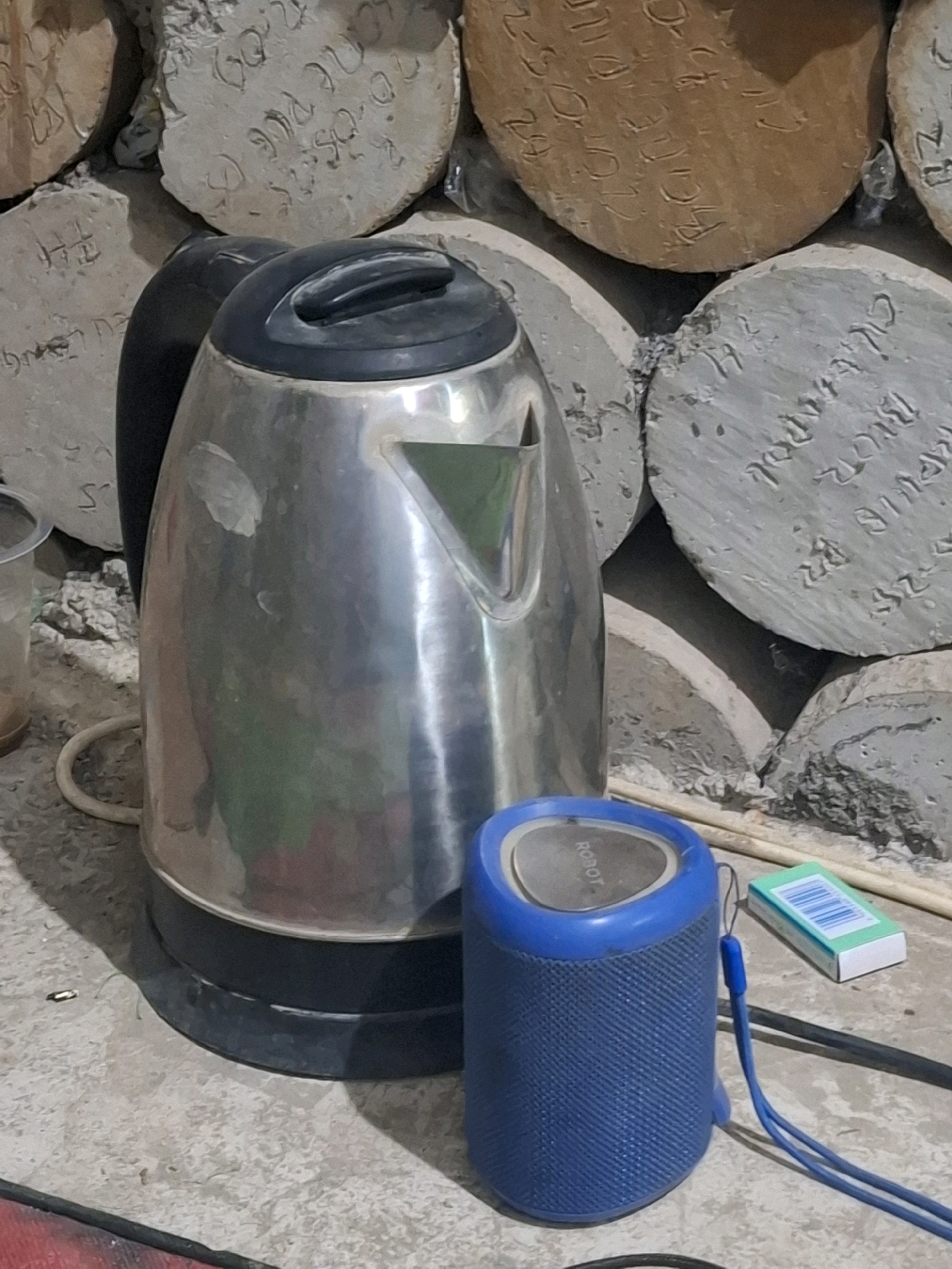 A silver electric kettle sits on a concrete floor next to a blue cylindrical speaker. In the background, stacked round concrete objects with engraved writing are visible. A small green and white box, possibly of cigarettes, lies to the right of the speaker. The scene appears to be outdoors, with a rough, textured surface dominating the foreground and background. The lighting suggests it is daytime, with shadows cast on the floor. The overall impression is one of an unkempt, possibly rural or construction-adjacent environment. The specific mention of Kutasirna, Indonesia, suggests this scene is located there.