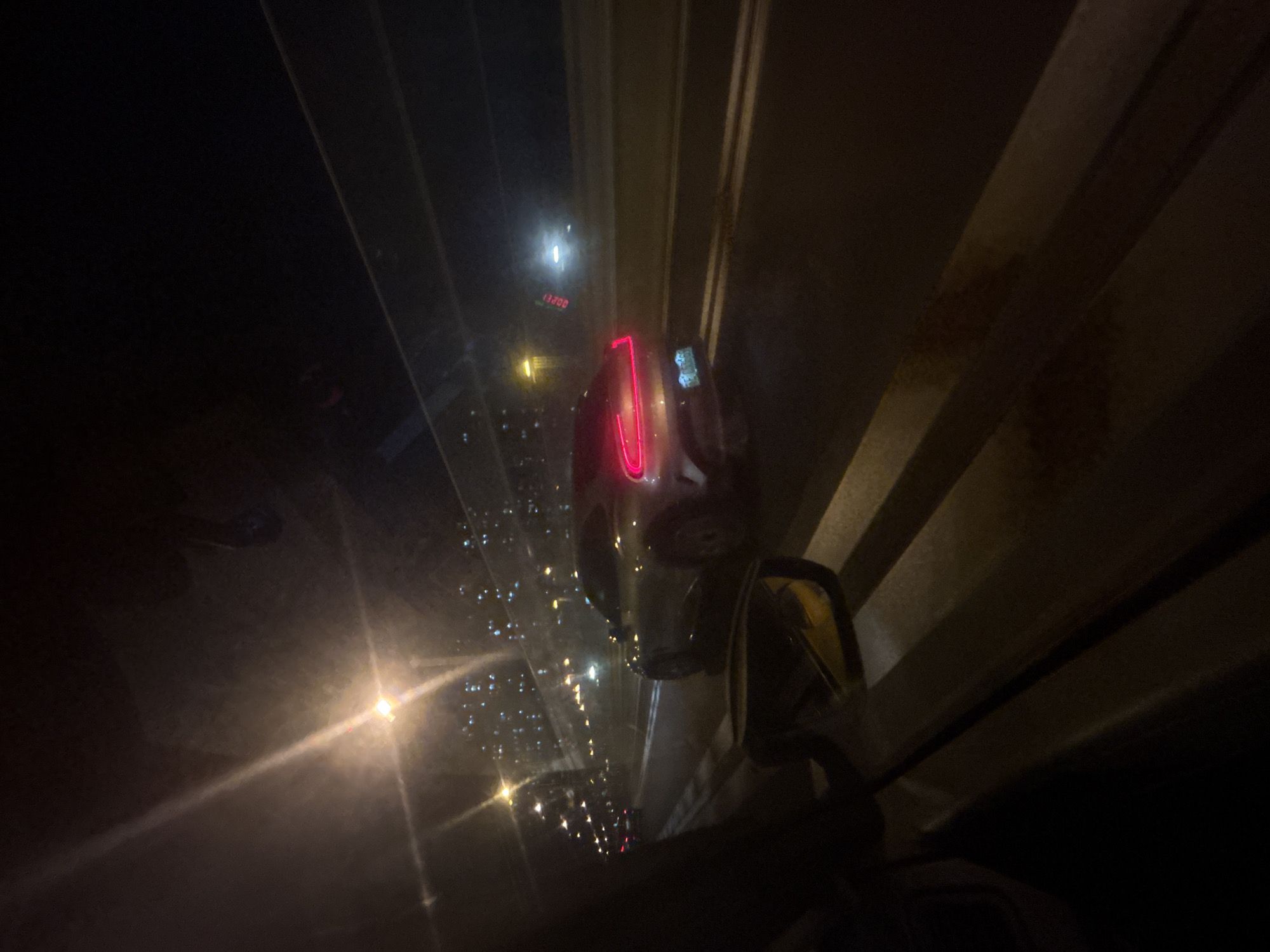 The image depicts a nighttime scene from within a vehicle, looking out at a highway. A silver sedan with its taillights illuminated in red is the primary subject, driving in the right lane ahead. The vehicle appears to be a Dodge Charger. Other vehicles and streetlights are visible in the background, along with blurred lights that suggest a city skyline, possibly in Suzhou Industrial Park, China. The view is partially obstructed by the interior of the vehicle, including a side mirror and parts of the dashboard and window. The overall lighting is dim, with prominent light streaks from streetlights and vehicle lights. The road surface appears wet, reflecting the lights.