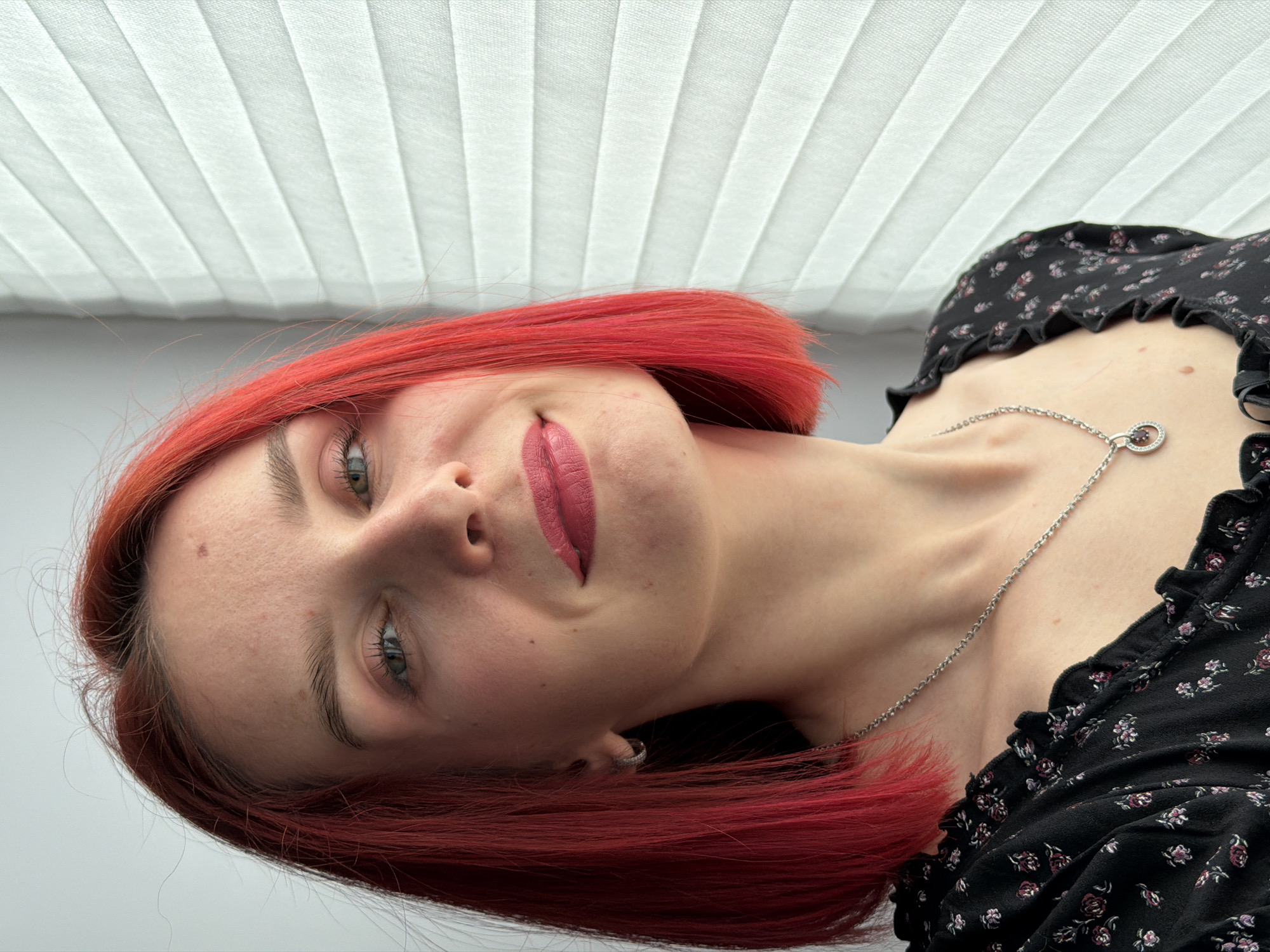 A medium-close-up photograph depicts a fair-skinned woman with dyed red hair. She is wearing a black, floral-patterned top and a silver chain necklace with a circular pendant. Her makeup includes light pink lipstick and visible eyebrow styling. She is smiling subtly. The background consists of white blinds and a white wall. The image is taken at an oblique angle, with the subject's head tilted. There is no verifiable information to suggest the location is in the city of Chaikovsky, Russia.