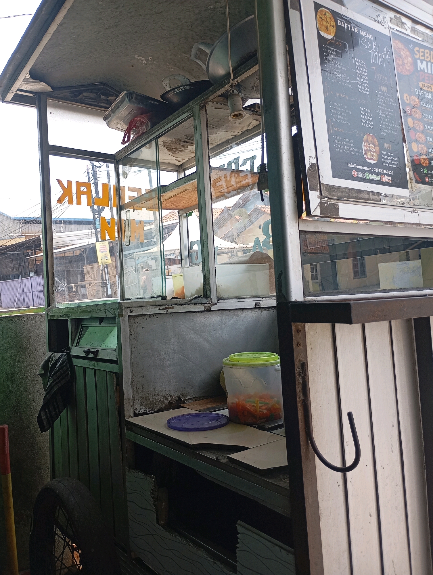 The image depicts a food cart in Babelan, Indonesia. The cart is made of metal and glass, with a menu board displayed on the right side. The menu board lists various food items and their prices in Indonesian Rupiah. Inside the cart, there are containers for food, and a plastic bucket with a green lid is visible on the counter. A bicycle wheel is attached to the bottom of the cart on the left side. The background shows buildings and other structures, suggesting an urban or semi-urban environment. The overall impression is of a small, functional food business operating on a street.
