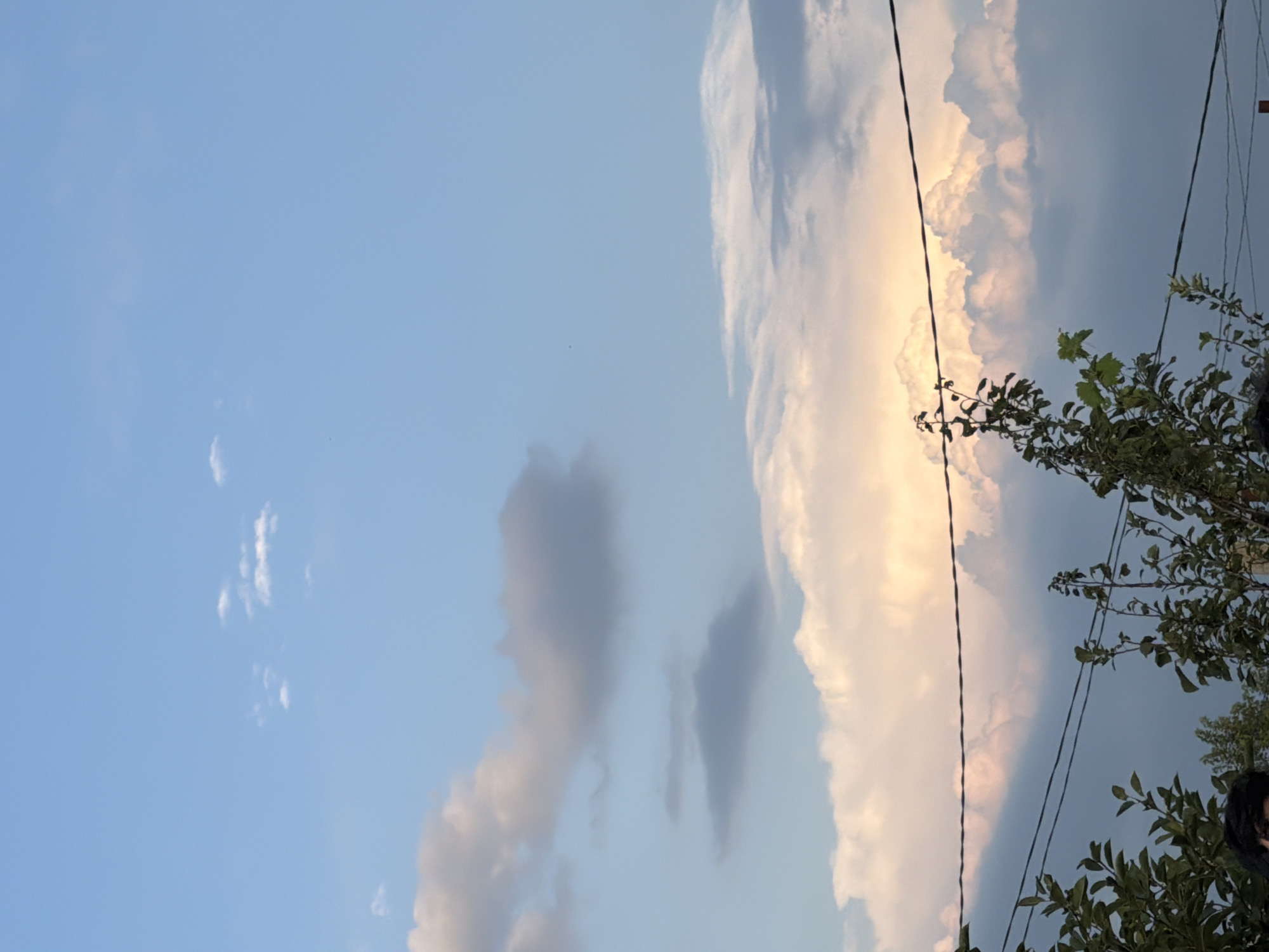 An outdoor, low-angle, vertical shot depicts a partially cloudy sky over Tbilisi, Georgia. The sky transitions from a lighter blue at the top to a slightly darker, grayish blue towards the bottom. Several clouds are scattered across the sky. In the upper portion, small, wispy white clouds are visible. Mid-sky, a larger, gray cloud is present, with a smaller, darker gray cloud situated below it. Near the bottom of the sky, a large, voluminous cloud formation is illuminated from behind by a golden-orange light, suggesting the sun is low on the horizon. In the foreground, the tops of green trees with dense foliage are visible. Two dark power lines stretch horizontally across the frame, one above the tree line and another slightly lower. A person's dark hair is partially visible in the lower left corner of the image, peeking out from behind the foliage. The overall scene suggests an evening sky, possibly during sunset.