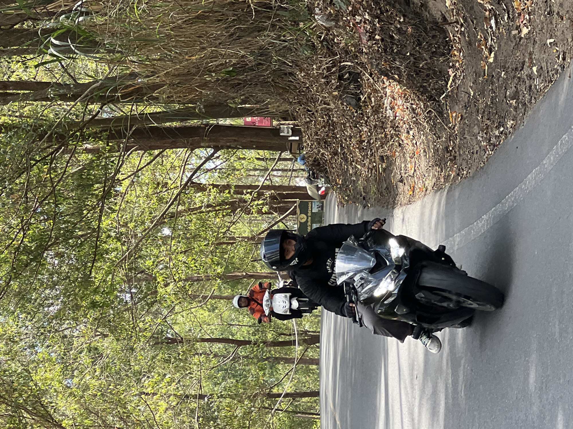 In Kintamani, Indonesia, two individuals are riding motorcycles on a paved road winding through a forested area. The motorcycle in the foreground is black with its headlights illuminated and is ridden by a person wearing a black jacket, dark pants, gloves, and a black helmet. The rider's left leg is extended slightly. Behind them, a white scooter is ridden by a person wearing an orange jacket and a white helmet. The road is bordered by dry brush and fallen leaves on the right, and tall trees with green foliage on both sides, creating a dappled light effect. Several signs are visible on the right side of the road, along with piles of debris and trash. The environment appears natural and somewhat untended.