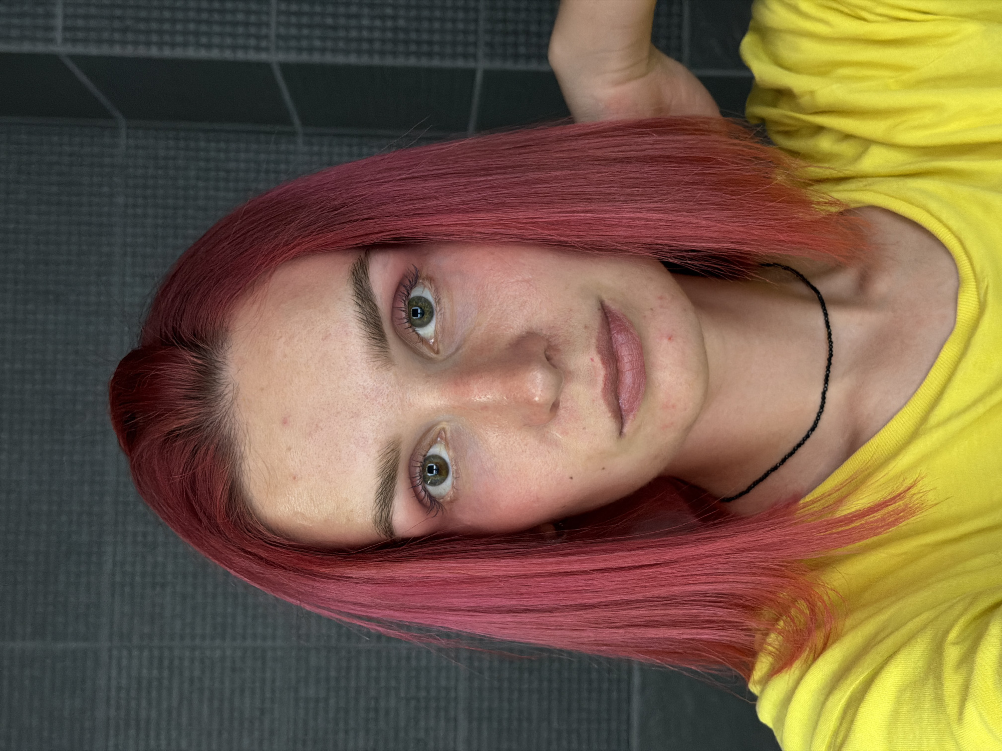 A young woman with bright pink hair and a yellow t-shirt is shown from the chest up. She is looking directly at the camera with a neutral expression. Her right hand is resting behind her head. The background appears to be a tiled wall. The image does not provide any visual information to ascertain the location in the city of Chaikovsky, Russia.