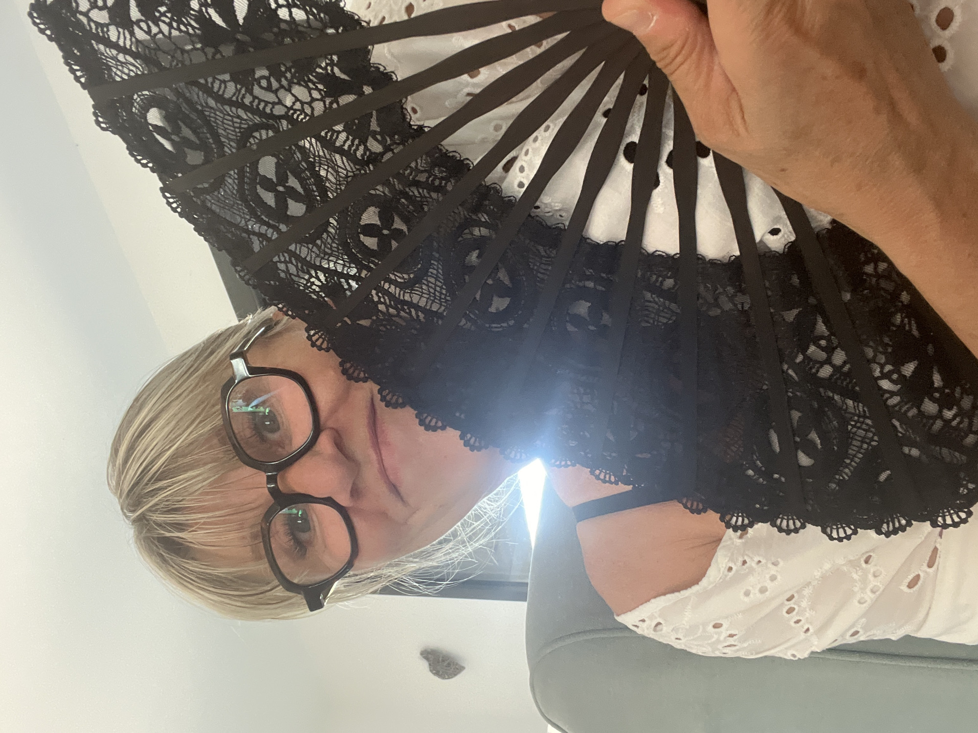 The image shows a woman indoors, holding a black lace fan. She is wearing glasses and a white eyelet top. The woman is looking towards the camera with a slight smile. The background suggests an indoor setting, possibly a living room, with a chair and a wall visible. There is no indication of the specific location in Chambœuf, France, or any contextual information about the scene.