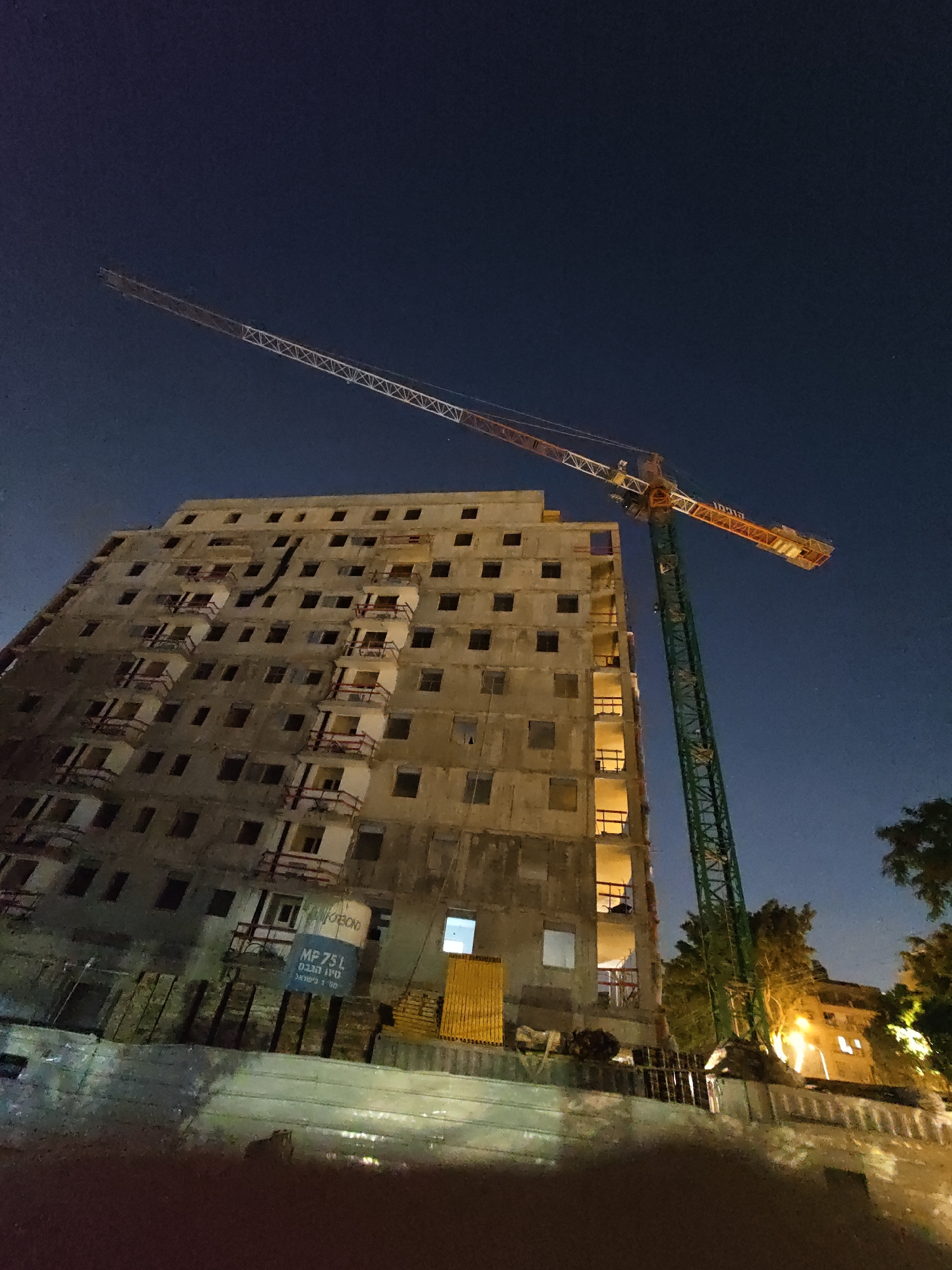 An exterior, low-angle, wide shot captures a construction site at night in Petah Tikva, Israel. A tall, green tower crane is prominently positioned to the right of a multi-story, unfinished building. The building, constructed from concrete, has numerous windows, some of which are illuminated, suggesting interior lighting. Balconies with red railings are visible on several floors. In the lower center of the image, a blue sign with white lettering is affixed to the building, displaying "MP 75L" and text in Hebrew that translates to "Made in Israel." Various construction materials, including wooden planks and metal fencing, are scattered around the base of the building. The sky is a deep blue, indicating it is nighttime, and a few trees are visible on the right side of the frame, with one illuminated by a light source in the background. The overall scene suggests ongoing construction work.