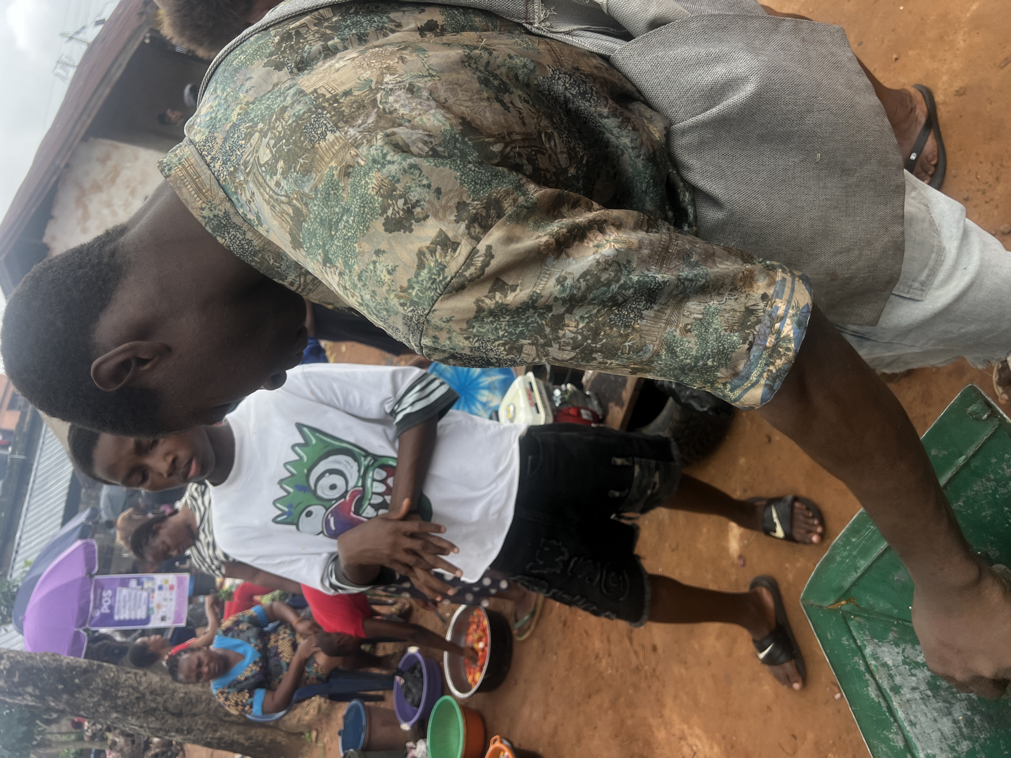 In Nkpor, Nigeria, a busy market scene unfolds. In the foreground, a young man wearing a white t-shirt with a cartoon graphic and black shorts with distressed details, stands with his hands clasped. He is wearing black flip-flops. To his right, another person, partially visible, wears a light-colored shirt with a detailed, nature-themed print and light-wash jeans, along with black flip-flops. A gray bag is slung over their shoulder.
The ground is made of reddish-brown soil, indicative of the market's location. To the left of the young man, several large bowls are arranged. One green bowl is partially visible in the bottom left corner. Adjacent to it is a larger orange bowl. Beyond that, a black bowl is filled with chopped red tomatoes. Further left, a purple bowl is also visible, and behind it, a blue plastic container.
In the background, a woman in a patterned blue and yellow dress sits on a blue chair, her hands resting on her lap. Another woman with dark hair is visible behind her, wearing a striped shirt. A purple umbrella with a "POS" sign attached is set up in the background. Other market-goers and stalls are present, contributing to the lively atmosphere of the outdoor market. The overall scene suggests a vibrant and active commercial hub.
