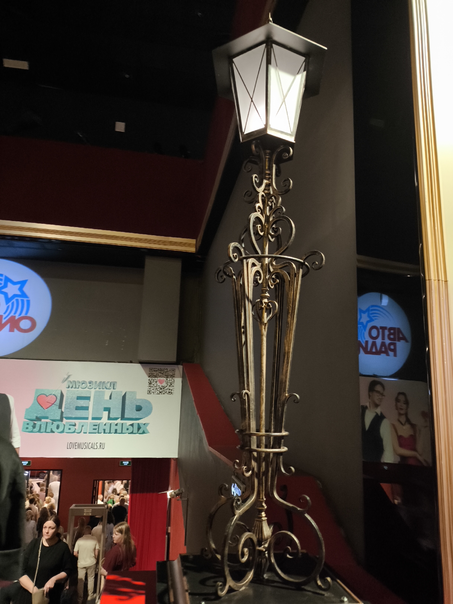 In an indoor setting with a dark ceiling and maroon-colored walls, an ornate, antique-style streetlamp with a white, illuminated lantern stands prominently in the foreground, angled slightly to the right. The lamp's base is intricate, featuring decorative metalwork. To the left of the lamp, a large sign is visible, displaying the words "МУЗЫКА" (music) and "ДЕНЬ ВЛЮБЛЕННЫХ" (day of lovers) with a red heart symbol. Below this, the website "LOVEMUSICALS.RU" is displayed. Further left, a circular light projection with a blue star and Cyrillic text is partially visible. To the right of the streetlamp, a large, dark, reflective surface acts as a mirror or screen, showing a reflection of a couple, a man in a suit and a woman in a red dress, along with a circular light projection that appears to be the same as the one on the left, but in reverse. In the background, an entrance to a venue is visible, with many people entering, suggesting a public event or performance. The lighting is dim, with focused illumination on the streetlamp and the signs.