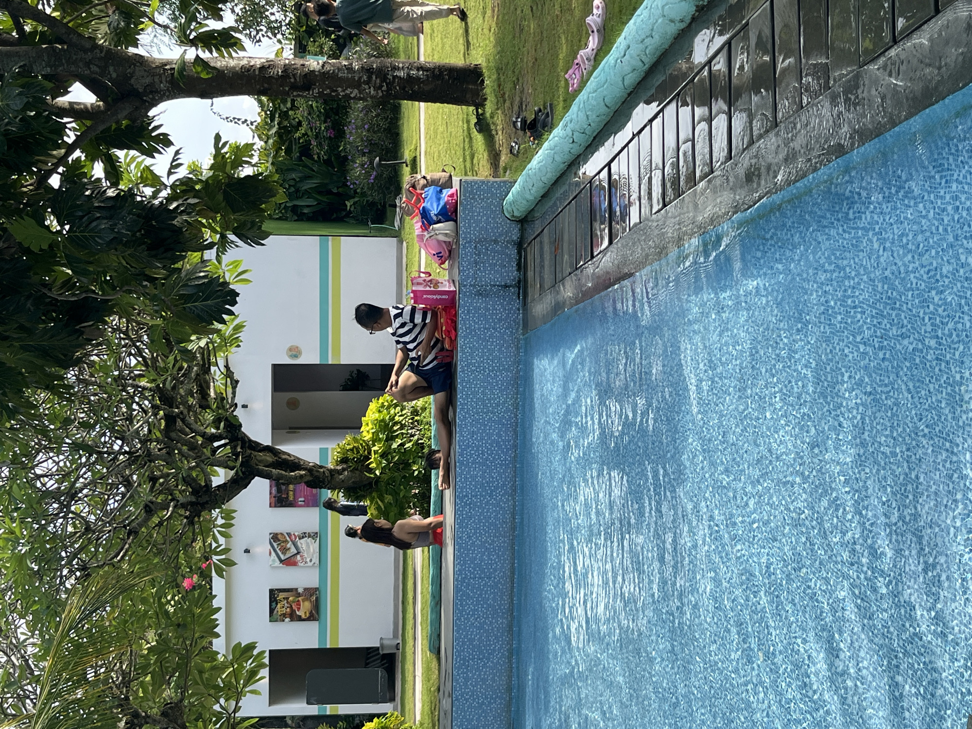The image depicts a poolside scene at what appears to be a resort or public pool facility in Kuta Selatan, Indonesia. The pool itself is a vibrant blue, tiled with a mosaic pattern. The edge of the pool is lined with a dark gray coping, and beyond that, a wooden deck extends outwards, with several pairs of footwear scattered on its surface. A bright teal pool noodle runs along the side of the deck.
In the background, a white building with a green and yellow striped band runs horizontally across its facade. Several framed pictures are mounted on the wall of the building. Adjacent to the building is a lush green lawn dotted with trees and tropical foliage.
Several individuals are present. In the foreground, a person wearing a striped shirt and dark shorts is reclining on the pool edge, appearing to relax. To their left, a person in a red swimsuit is sitting on the edge, facing away from the camera. Further left, a person in dark clothing is also seated. In the background, another person in a light-colored outfit is walking on the grass.
A collection of bags, including a pink one, a blue one, and several brown and beige bags, are placed on the pool's edge near the person in the striped shirt. Several pairs of flip-flops are visible on the grass and near the pool edge, including a pair of pink Crocs. The scene suggests a leisurely day spent outdoors, likely in a warm climate given the attire and environment.