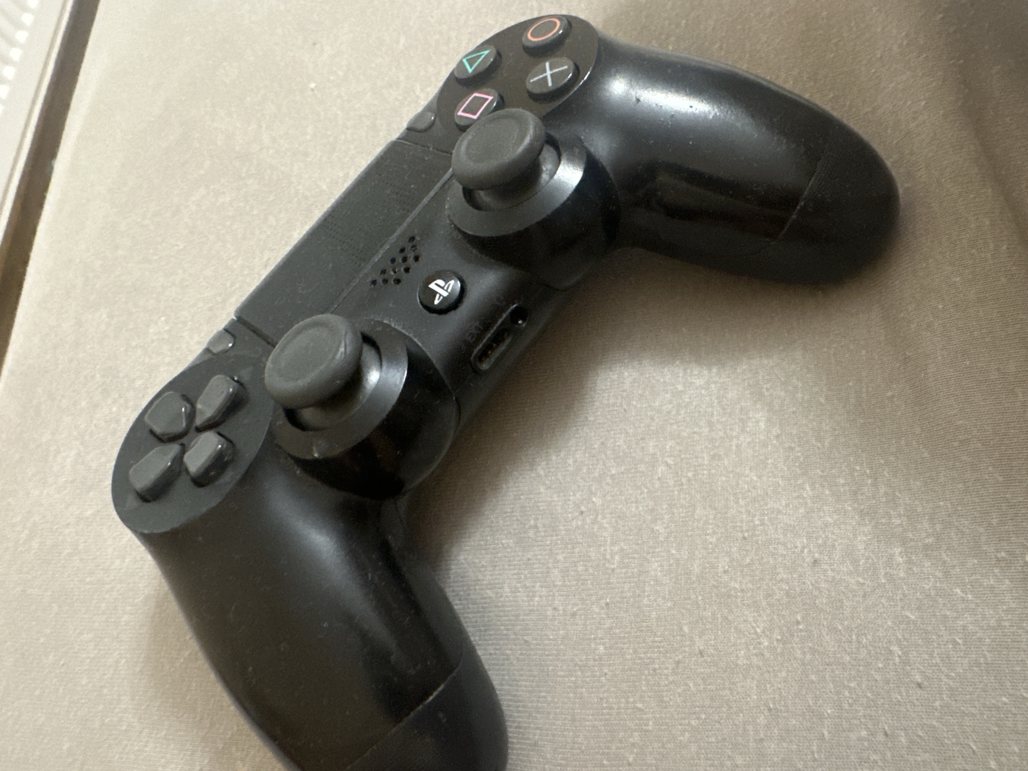 A black PlayStation 4 (PS4) DualShock 4 controller is the primary subject, resting diagonally on a light beige, textured surface. The controller is positioned with its right side angled away and slightly upwards, showcasing its upper-right half prominently.
The controller is a dark black color and exhibits signs of use, including some dust and lint on its surface. Key features visible on the controller include:
- The right analog stick, situated towards the upper-right area.
- Four distinct face buttons: triangle, circle, X (cross), and square symbols, which are etched in white or light gray on the black buttons.
- Below the face buttons and to the left of the right analog stick, a PlayStation (PS) logo button is clearly visible.
- A speaker grille is located to the left of the PS button.
- A micro-USB charging port is present on the front edge of the controller, below the PS button.
- On the left side of the controller, partially visible, are the four directional buttons of the D-pad, and a portion of the left analog stick.
The surface underneath the controller appears to be a fabric, possibly upholstery or carpet, characterized by a fine, horizontal texture and visible pilling (small fabric balls), indicating wear. In the top-left background, a thin, vertical, light-colored edge, possibly part of a wall or furniture, is faintly discernible. The scene is indoors. No specific time of day or outdoor weather cues are present.