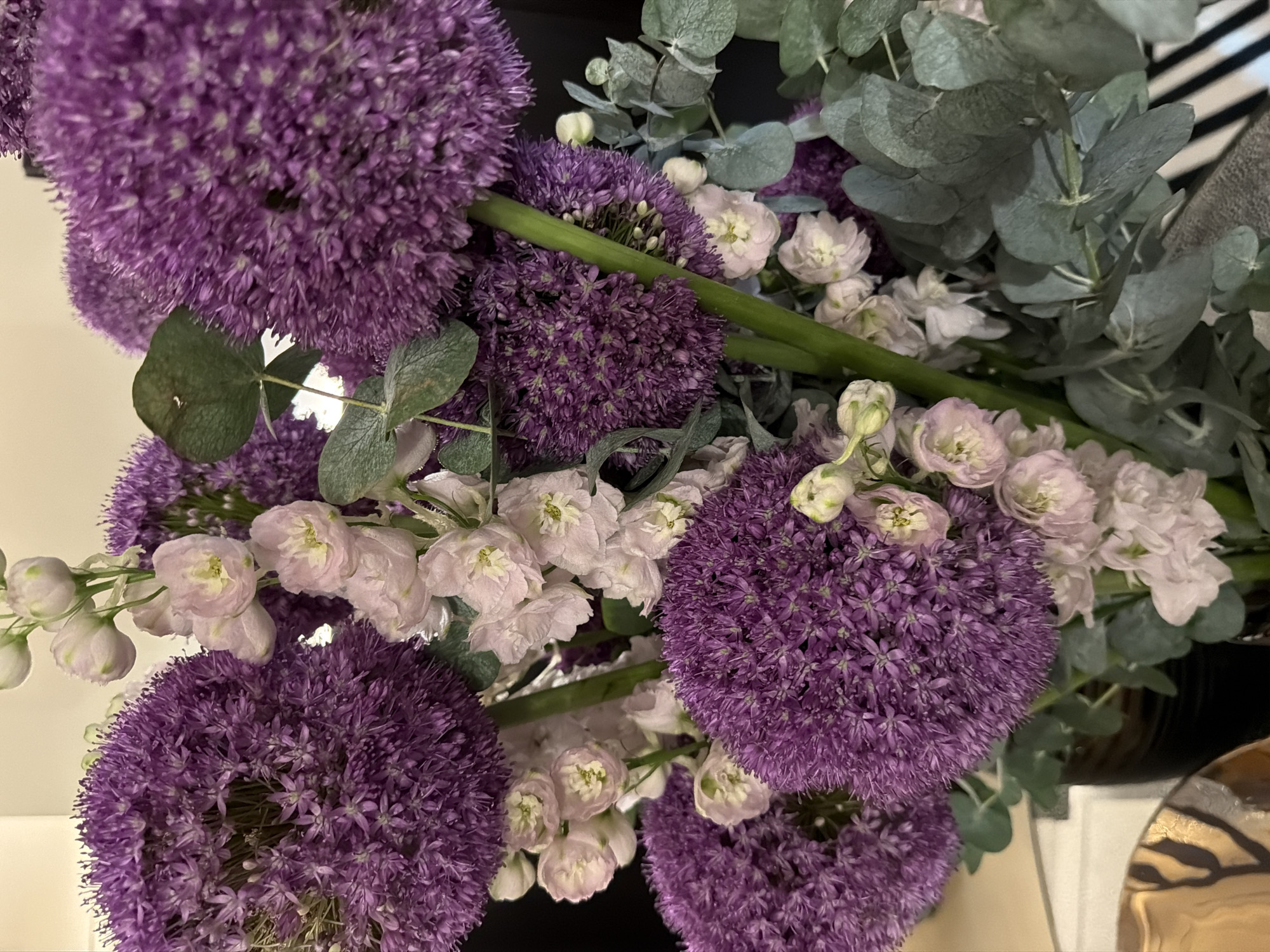 This image shows a close-up of a floral arrangement. The arrangement features several large, spherical purple allium flowers, interspersed with smaller, pale pink delphinium flowers. Eucalyptus leaves, with their characteristic silvery-green hue, are also present, adding texture and volume. The flowers are arranged in a dark vase, which is partially visible at the bottom left of the frame. The background is soft and out of focus, appearing to be a neutral-toned wall and possibly a glimpse of a decorative object. There are no people or discernible actions taking place. The lighting is soft, highlighting the textures and colors of the flowers. The location in Moscow, Russia is not determinable from the visual information provided in the image.