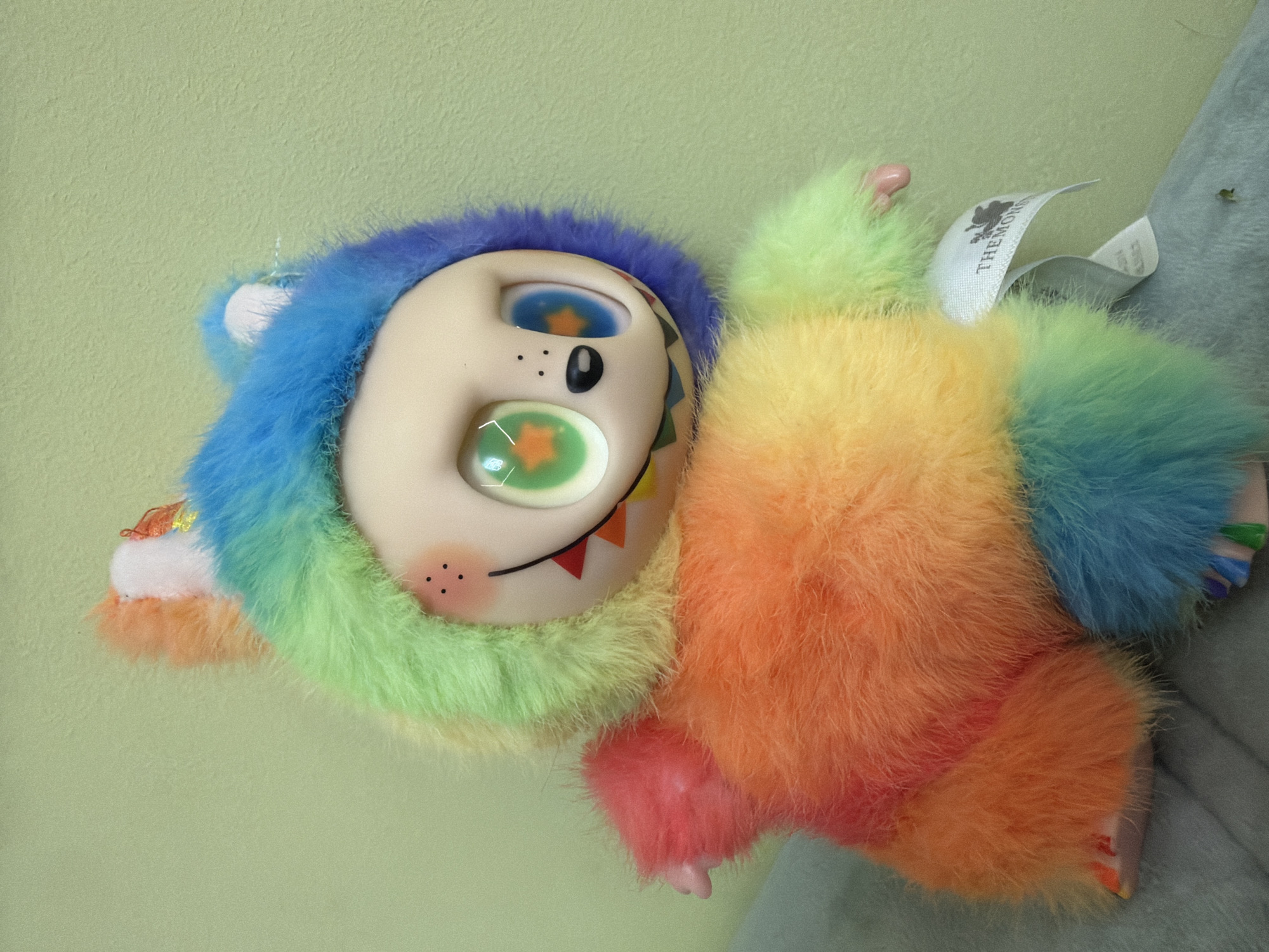 The image captures a vibrant, rainbow-colored furry plush toy, depicted indoors against a muted light green wall. This playful creature, located in Chaikovsky, Russia, features a large, smooth, light-toned face with a whimsical and cheerful expression. Its distinct, recessed eyes are noteworthy: one is blue with an orange star pupil, and the other is green, also with an orange star. A small dotted nose and rosy cheeks, indicated by clusters of red dots, are visible on its face. A wide, upward-curving smile reveals small, triangular, pointed elements, adding to its friendly yet slightly mischievous appearance.
The toy's body is covered in shaggy, soft fur that transitions through a spectrum of colors including blue, green, yellow, orange, and pink, resembling a rainbow. It appears to be resting on a soft, grey fabric surface on its right side. A white fabric tag is partially visible near the toy's back, displaying the legible text "THEMON" and the numbers "32024", likely indicating a brand or product identification. The lighting suggests an indoor setting, and no specific time of day or external weather conditions are discernible. The overall impression is one of bright colors and a charming, fantastical character.