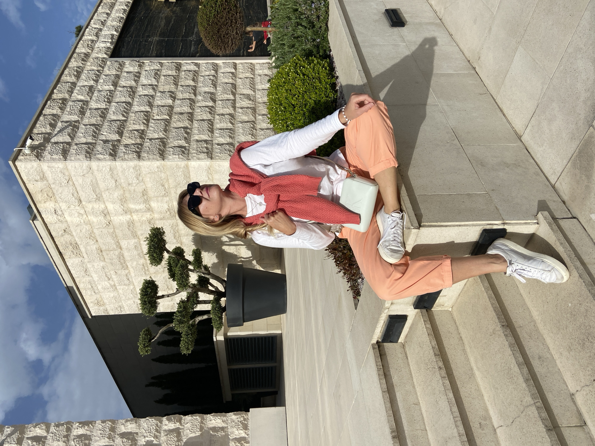 A woman is sitting on a stone staircase outside a modern building in Budva, Montenegro. The building has a facade of textured stone and black panels. She is wearing a white long-sleeved shirt, peach-colored pants, a coral sweater draped over her shoulders, and white sneakers. She also has sunglasses on her head and is holding a light-colored purse. A potted tree sits beside the building.