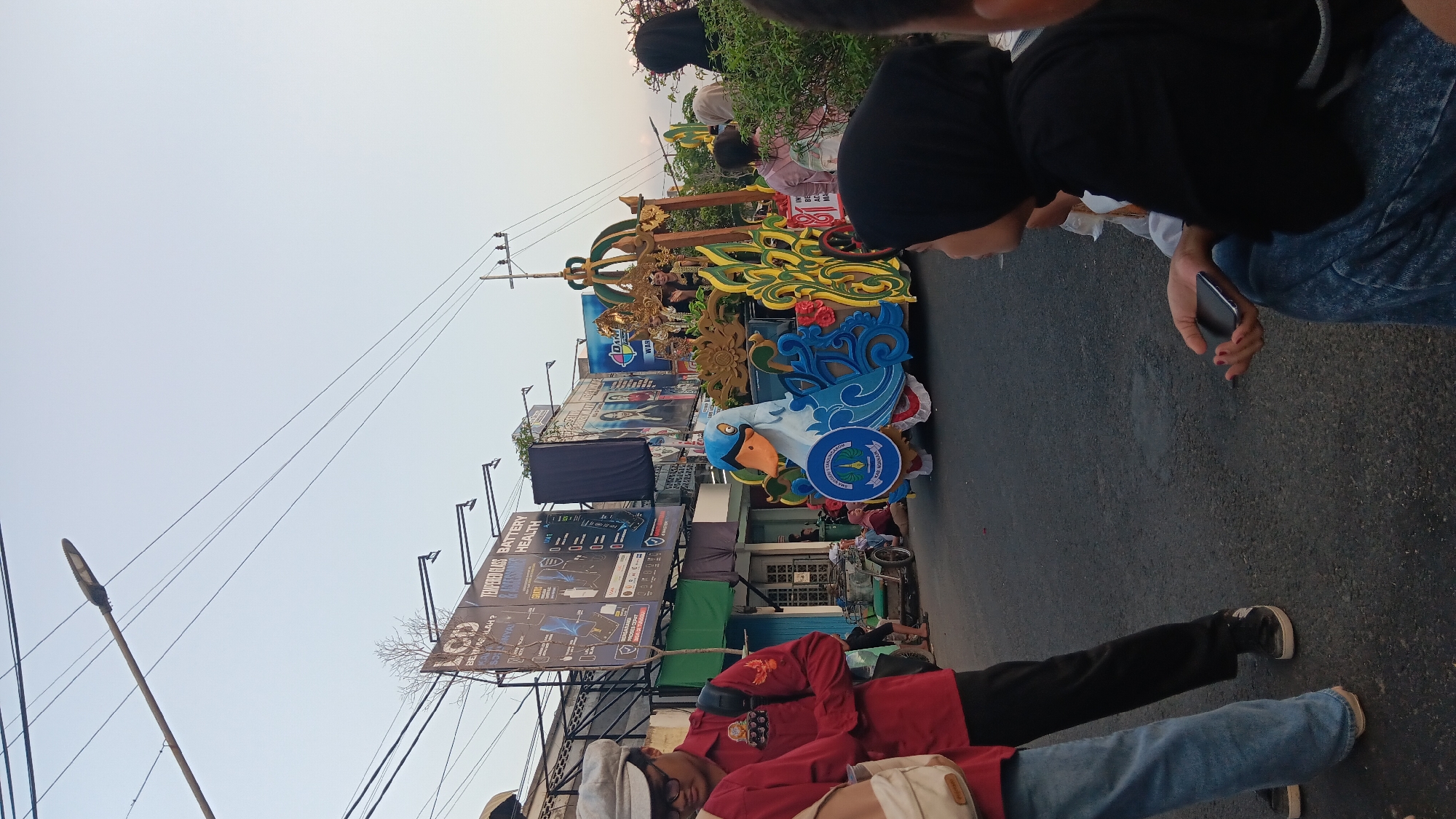 The image depicts a street scene in Tanjung Anom, Indonesia, possibly during a parade or festival. A decorative float featuring a large blue swan with an orange beak and an intricate golden and green ornamental structure is positioned on the right side of the road. Several people are visible, including individuals in the foreground wearing casual clothing and a person in a red shirt with a backpack on the left. A few individuals are seated on the side of the road, observing the event. In the background, buildings with advertisements and utility poles with overhead wires line the street, indicating an urban environment. A small motorized tricycle is parked on the left side of the road. The sky is clear and appears to be late afternoon or early evening.