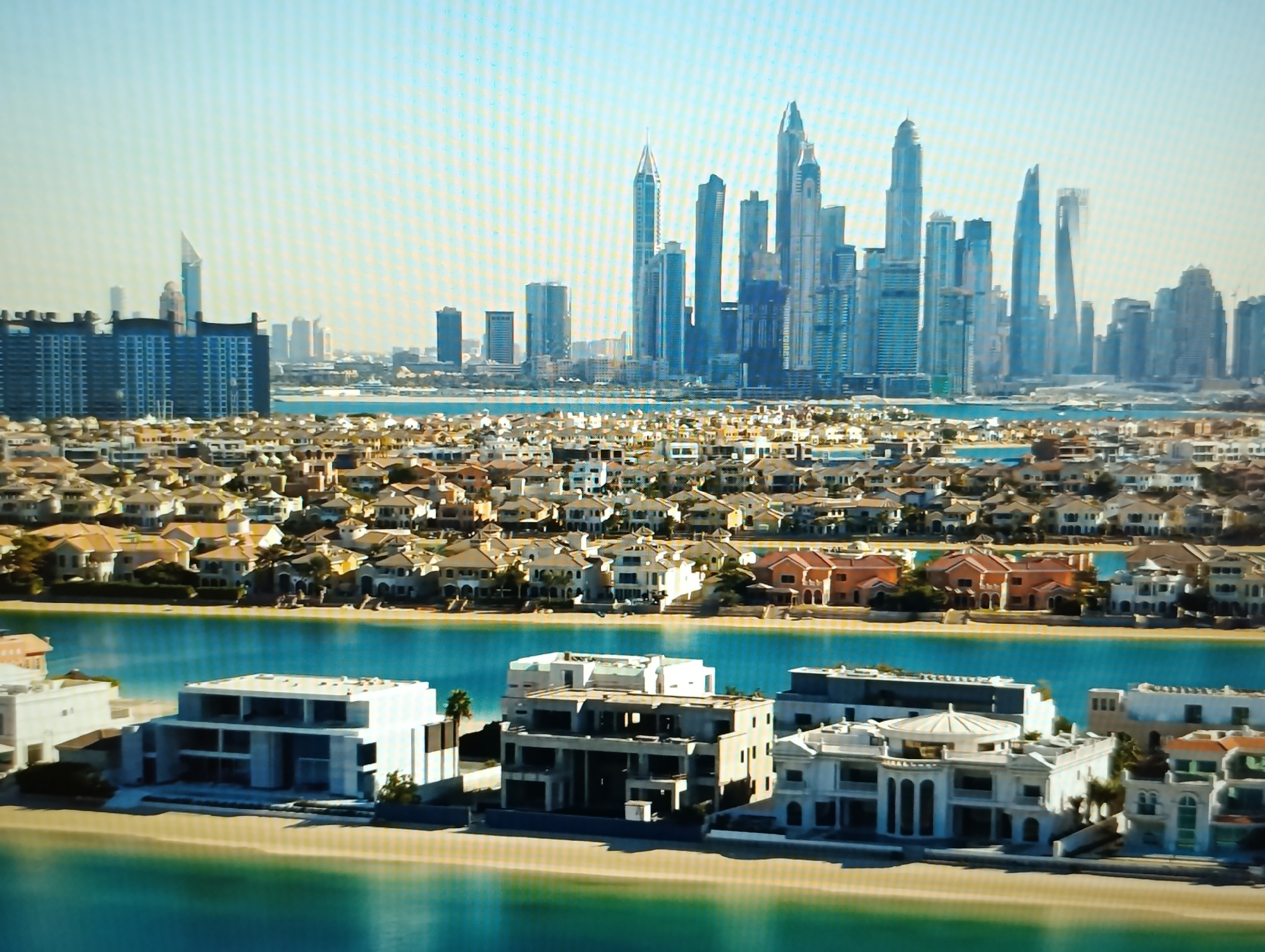 The image depicts a cityscape with a modern architectural style, dominated by a cluster of towering skyscrapers in the background, characteristic of Dubai. In the foreground, a waterfront area is visible, featuring several large, luxurious villas with contemporary designs. These villas are situated on man-made islands or peninsulas, bordered by tranquil blue water and sandy beaches. The overall scene conveys a sense of affluence and advanced urban development. The prompt's reference to Dubăsari town council, Moldova, is incongruous with the visual content of the image, which clearly shows a Dubai skyline.