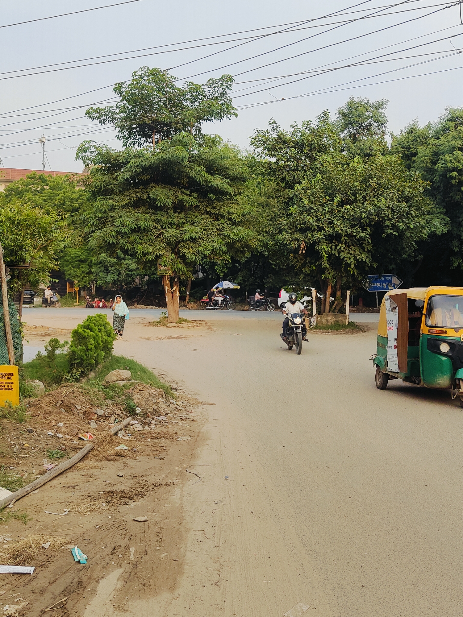 This image depicts a street scene in Gurgaon, India, with an emphasis on a busy intersection. In the foreground, a dirt path is visible, littered with debris including pieces of wood, straw, and what appears to be a plastic wrapper. A yellow sign with text is partially visible on the left side of the frame.
Further into the scene, a paved road curves into the distance. A woman in a light blue garment and a headscarf is walking on the side of the road, facing the camera. A few small green bushes are planted in a row to her left. In the background, several trees with dense green foliage line the street.
Several vehicles are present. A three-wheeled auto-rickshaw, painted green and yellow, is parked on the right side of the road, facing away from the camera. A man is riding a motorcycle towards the viewer. Further back, a few other motorcycles and a white car are visible. People are gathered in small groups near the intersection. A street sign with blue text is mounted on a pole. The sky is overcast, suggesting a cloudy or hazy day. The overall impression is one of an active, somewhat unkempt urban environment.