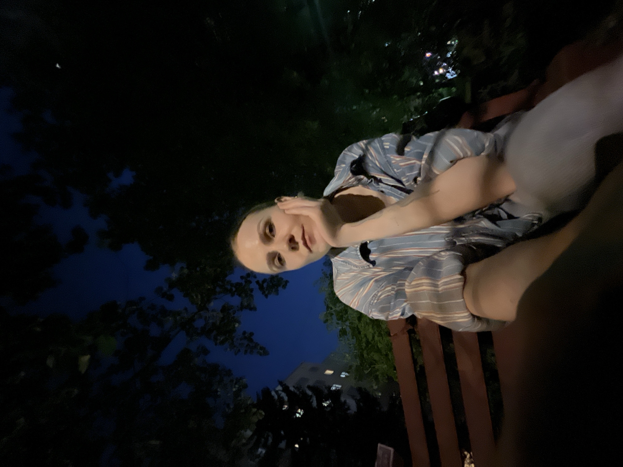A medium-shot of a person with light skin, resting their chin on their hand, wearing a blue and white striped shirt, and seated on a wooden bench. The background features a dark, possibly nighttime, environment with trees and a building illuminated by lights. The location is possibly Khimki, Russia.