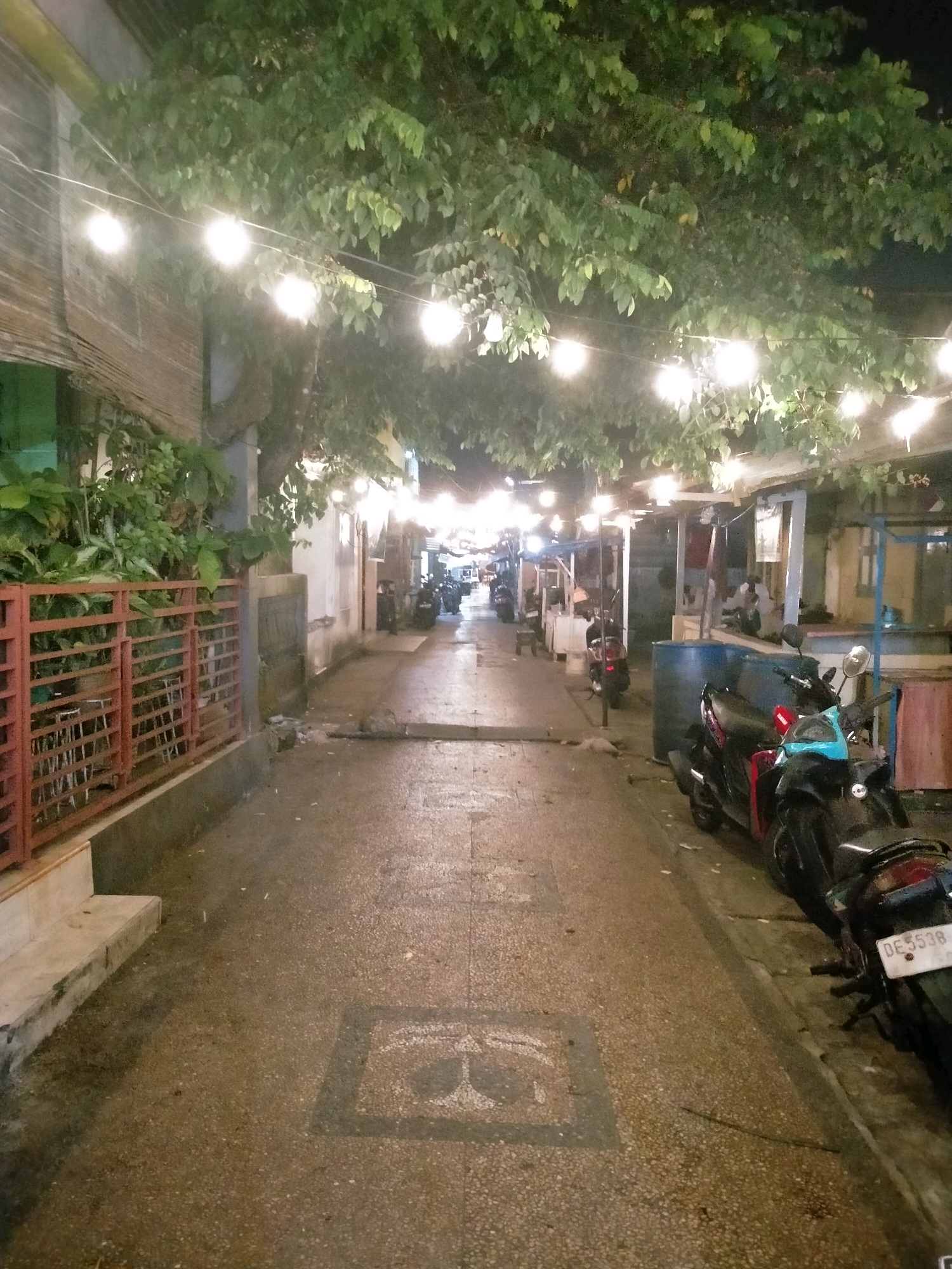 A narrow street in Ambon, Indonesia, is illuminated by strings of overhead lights. The street is paved with rough, light-colored gravel and asphalt. To the left, a red metal fence encloses an area with tables and chairs, suggesting an outdoor seating area for a cafe or restaurant. Lush green foliage hangs from trees overhead, and some vegetation grows behind the fence.
To the right of the street, a row of buildings lines the path, some appearing to be shops or food stalls. Several motorcycles are parked along the right side of the street. One motorcycle in the foreground, a dark color with red accents and a blue seat, has its license plate visible, reading "DE 5538".
The scene is depicted at night, with artificial lights casting shadows and highlighting the wet appearance of the pavement. The overall atmosphere is that of a bustling, lively street, possibly a marketplace or a dining district in Ambon.