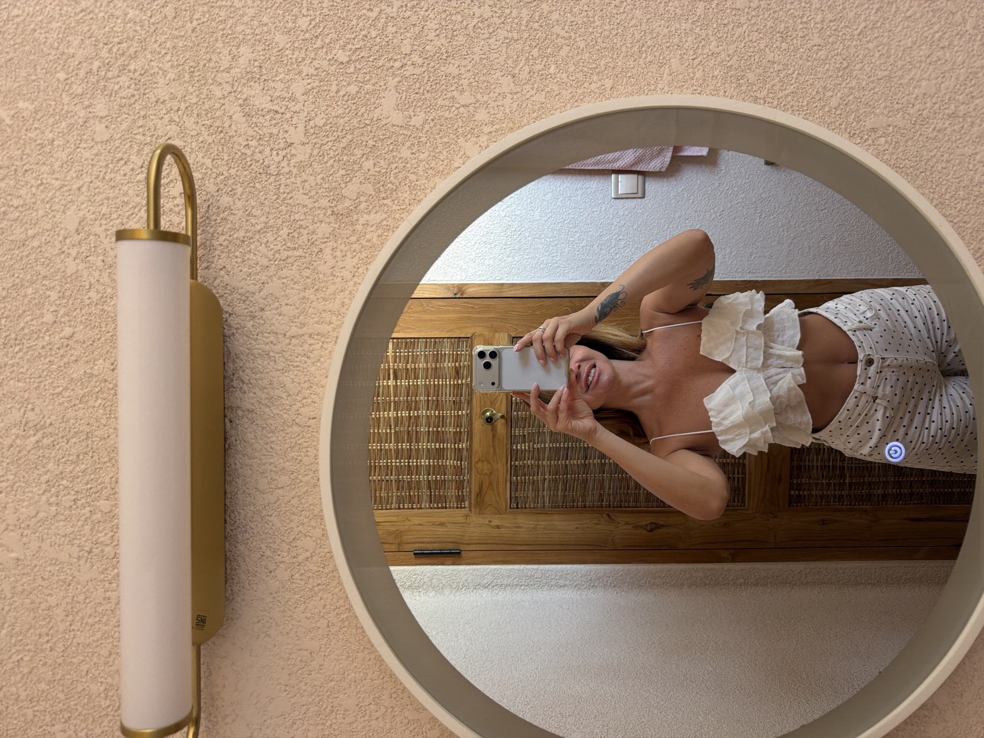 A woman takes a selfie in a round mirror. She is wearing a white ruffled crop top and white polka dot pants. Her phone is held up to take the photo. The mirror reflects a wooden door with woven panels. The wall behind the mirror is textured and peach-colored. A gold and white light fixture is mounted on the wall above the mirror.
