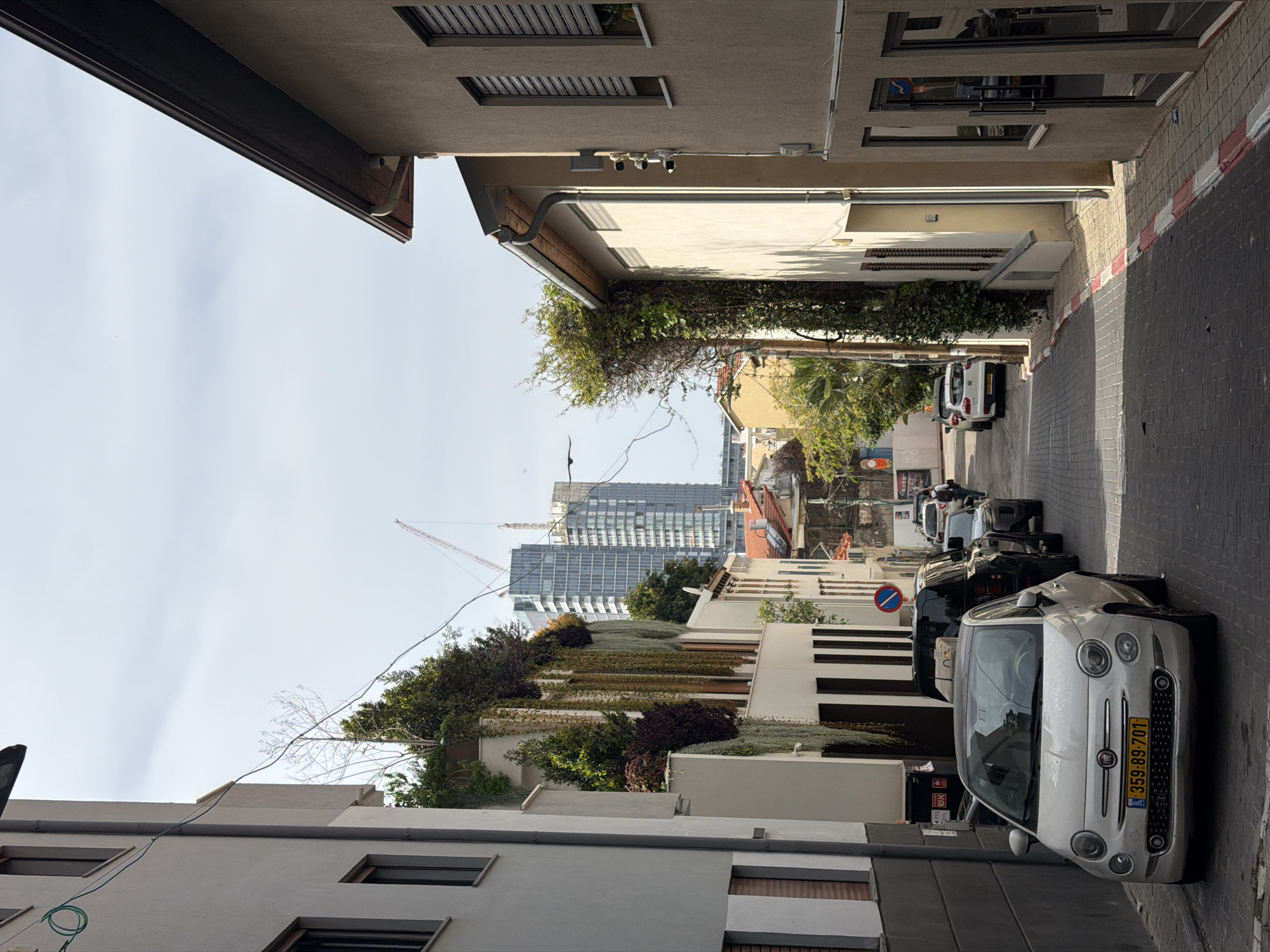 The image depicts a narrow street in Tel Aviv, Israel. The street is lined with buildings, some of which are covered in greenery. Several cars are parked along the street. In the distance, modern skyscrapers and construction cranes are visible, contrasting with the older architecture. The sky is partly cloudy. The perspective is looking down the street with building walls on each side.