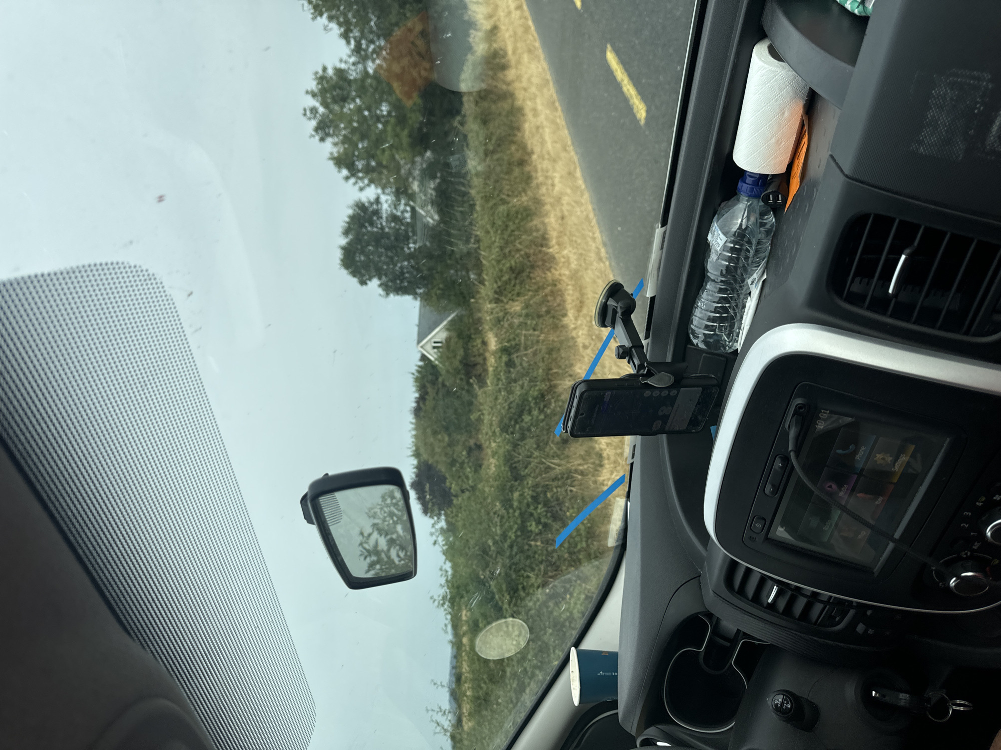 The image shows the interior of a vehicle driving on a rural road in Ireland. The perspective is from the driver's seat, looking out through the windshield. The dashboard is visible, with a car stereo, air vents, a phone mount with a phone, a cup, a water bottle, and a roll of toilet paper. The windshield has some smudges and a circular sticker. Outside the vehicle, the road is visible with yellow lines. Beyond the road are fields of dry grass and green bushes, with trees and two houses in the background. The sky is overcast. The time on the car stereo is 10:01.