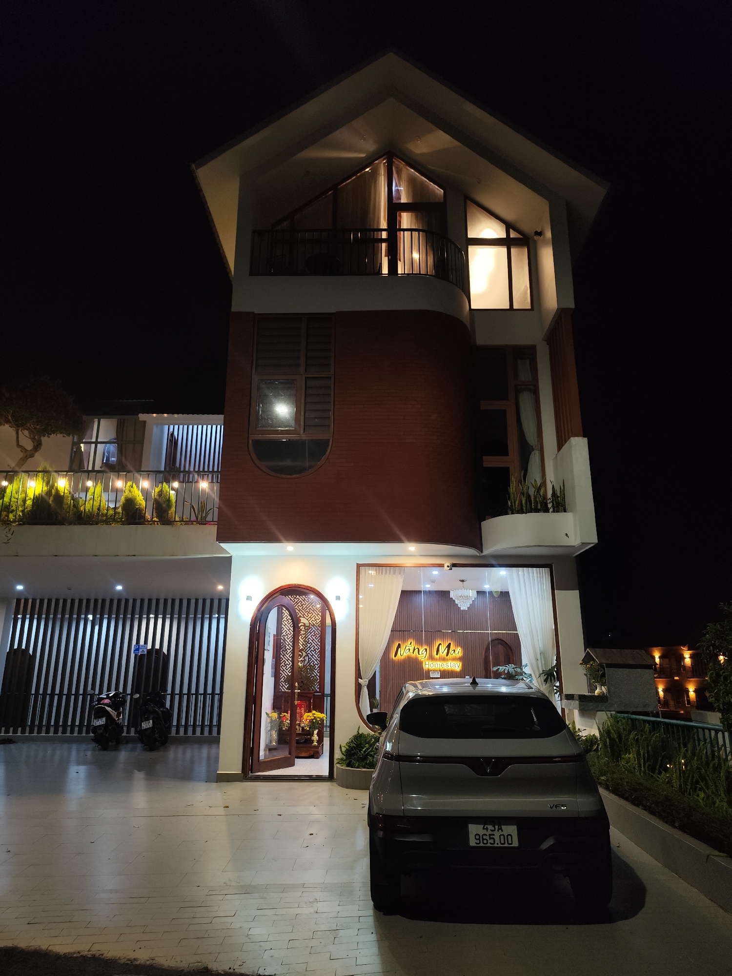 This is a nighttime exterior shot of a multi-story homestay named "Nàng Mai Homestay" located in Da Lat, Vietnam. The building features a modern architectural style with a combination of white, brick, and wooden elements. The facade is illuminated by exterior lights, creating a warm glow.
In the foreground, a white electric vehicle is parked on a paved driveway. The vehicle has a license plate reading "43A 965.00". To the left of the vehicle, two motorcycles are parked under a covered area with vertical blinds.
The entrance to the homestay is visible, with a large, ornate wooden door. Through the open door, an interior with decorative elements and flowers can be seen. White curtains hang on either side of the entrance, and a neon sign with the homestay's name is displayed.
The upper floors of the building have balconies and large windows, some of which are illuminated from within, revealing glimpses of the interior. There are also several planters with greenery on the balconies.
In the background, the dark night sky is visible, with some distant lights from other buildings. The overall scene depicts a well-lit and inviting homestay property at night.