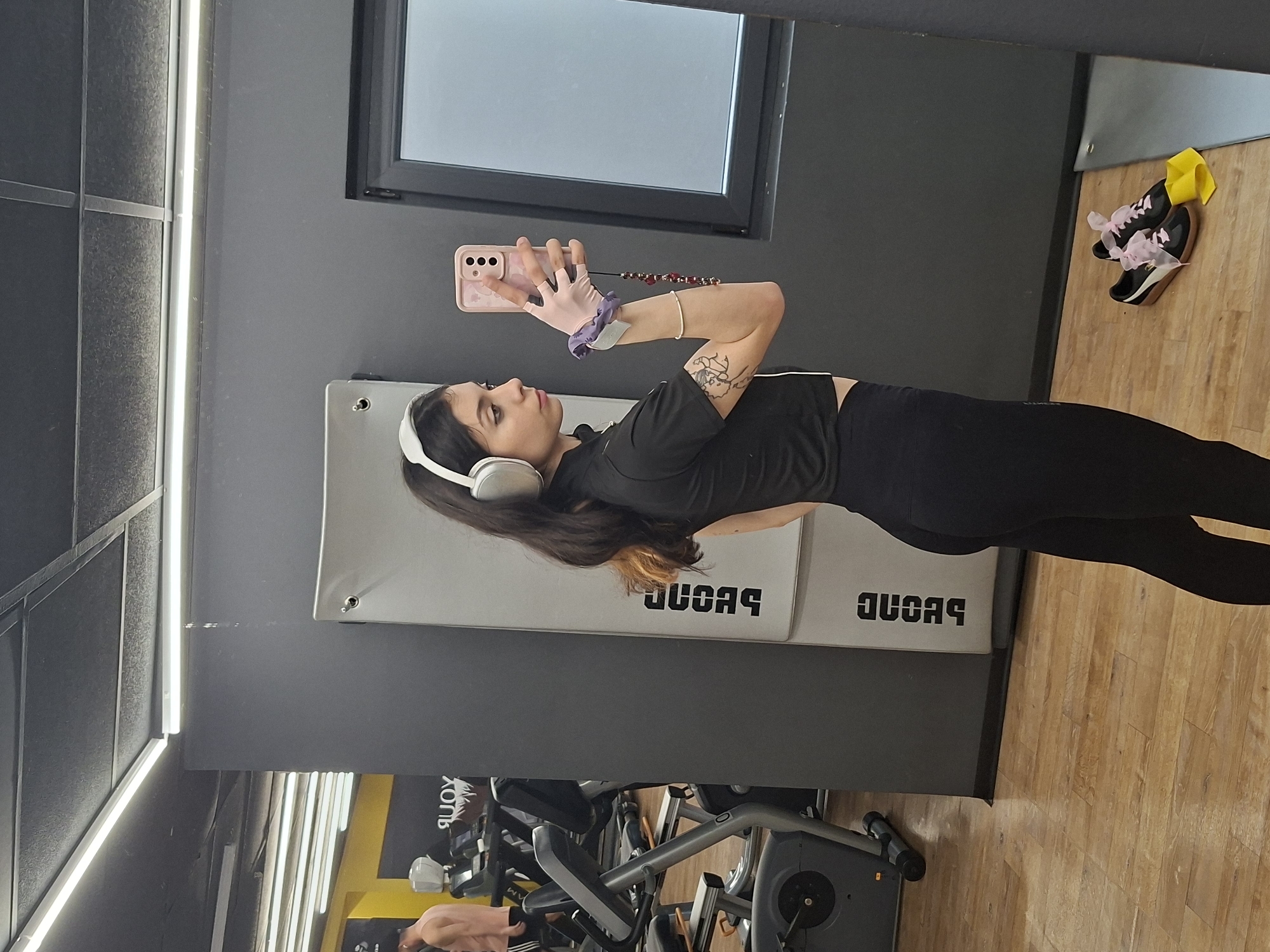 The image displays a mirror selfie taken by a young woman in a gym or fitness center, likely located in Sofia, Bulgaria. The woman, standing prominently in the frame, has long dark hair and is wearing a black short-sleeved top and black leggings. She is equipped with white over-ear headphones and pink fingerless fitness gloves. Her right arm, which bears a tattoo, holds a light pink smartphone with a patterned case and a red beaded strap, used to capture the reflection.
The setting features dark grey walls, a light wooden floor, and a large window with an indistinct view, suggesting an indoor daytime scene. Bright vertical fluorescent lights illuminate the space on the left. Behind the woman, there's a padded grey wall segment with text that appears as "DUCAR" when seen in the mirror, implying a brand name or gym identifier (perhaps "RACUD" or "DUCARD" in reality). Below this, various pieces of gym equipment are visible, including what looks like a stationary bike or similar apparatus. A faint reflection or background detail suggests another person is also in the gym, using equipment. On the wooden floor to the woman's right, a pair of black sneakers adorned with pink laces and decorative pink bows rests beside a folded yellow item, possibly a towel or accessory. The woman's expression is neutral, focused on taking the photograph.