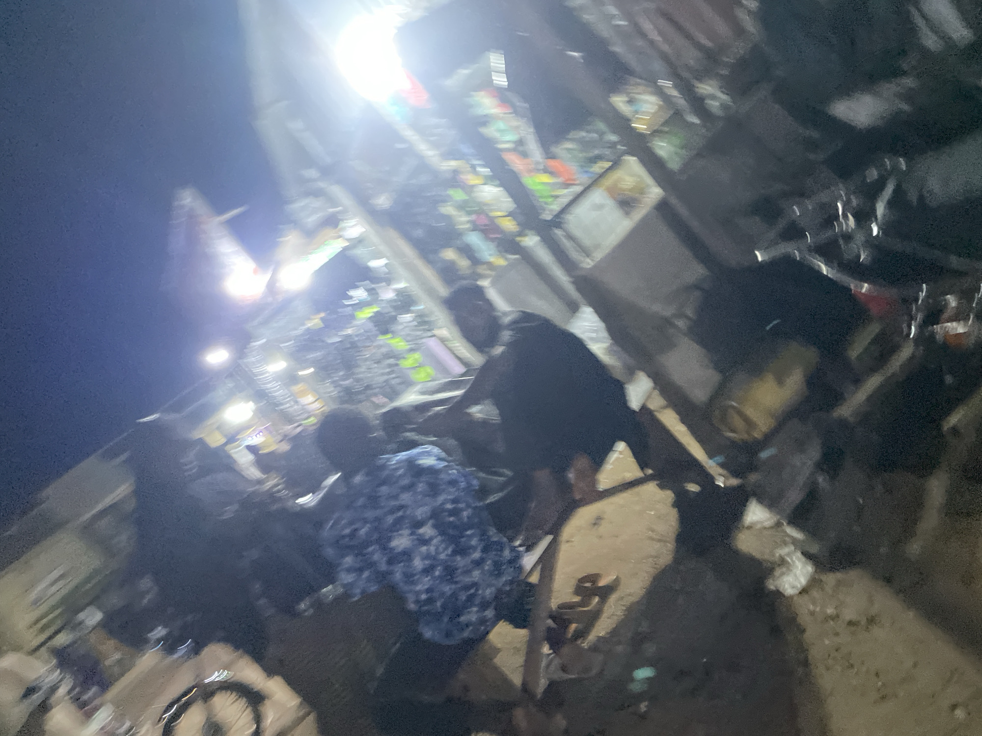 The image depicts a dimly lit, outdoor scene at night, possibly a street market or roadside stall in Lokoja, Nigeria. Several individuals are present, some of whom appear to be interacting with merchandise displayed on tables and shelves. The lighting is harsh and appears to be from artificial sources, creating bright glare and deep shadows that obscure details. The overall image is blurry and low resolution, making it difficult to ascertain precise actions or identify specific items. The environment appears somewhat cluttered, with visible debris on the ground. The orientation of the image is also tilted, adding to the disorienting nature of the scene.