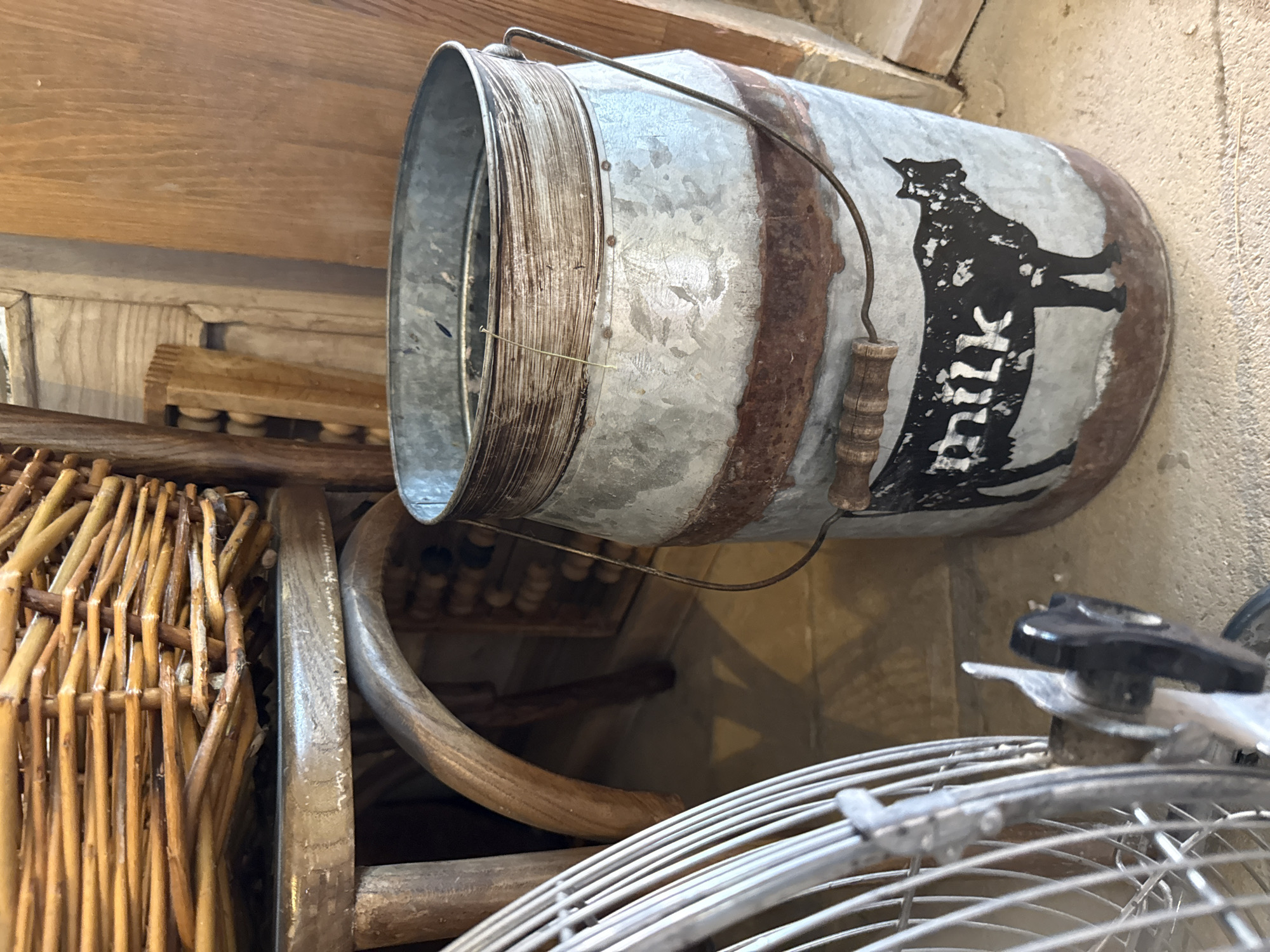 A vintage galvanized metal milk pail with a dark silhouette of a cow and the word "milk" painted on its side sits on a concrete floor. The pail has a wooden handle and a wire bail handle. Behind the pail, a wooden abacus and wooden planks are visible. To the left of the pail, a portion of a wire fan guard is in the foreground. The scene appears to be indoors, possibly in a rustic setting. The image does not provide any information about the city of Natukhaevskaya, Russia.