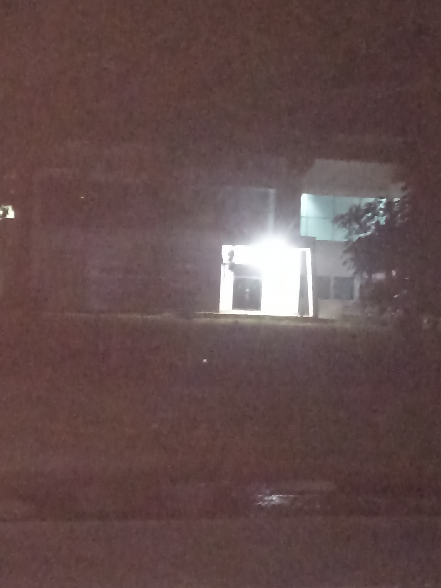 A nighttime, low-resolution, grainy, overexposed image captures a building exterior in Kampung Tengah, Indonesia. A brightly lit entrance or alcove dominates the central portion of the image, with a strong light source casting a white glow. The structure appears to be modern, with clean lines and possibly glass elements visible on upper floors. Trees are present to the right of the building. The foreground consists of dark, indistinct ground, possibly grass or pavement, with some wet patches or reflections visible. The overall scene is dim and somewhat obscured by the poor image quality and lighting conditions.