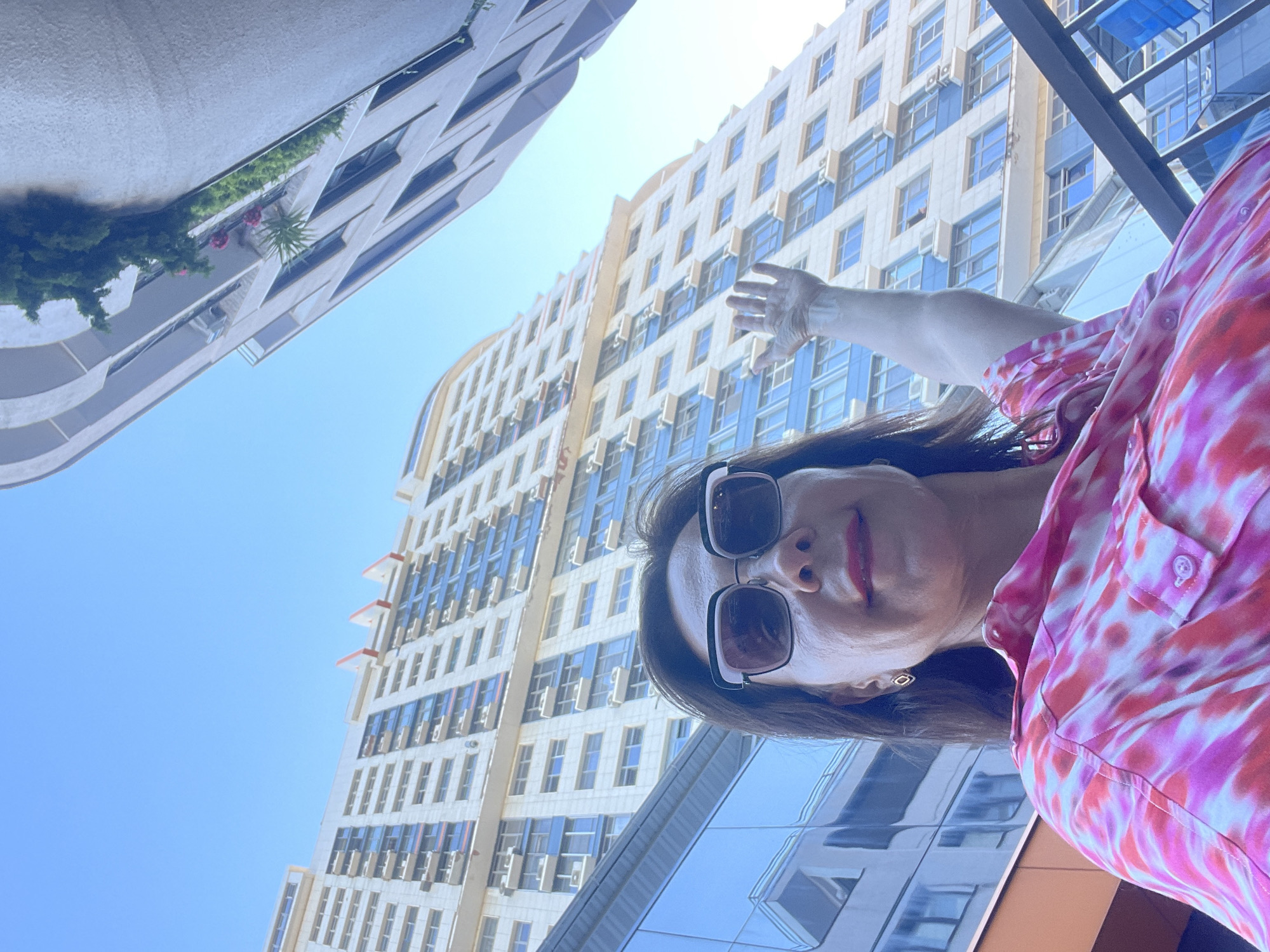 The image shows a woman in the foreground, taking a selfie. She is wearing sunglasses and a brightly colored pink and white tie-dye shirt. Her arm is outstretched, with her hand reaching towards the camera. Behind her, several modern buildings with many windows are visible, reaching high into a clear blue sky. The buildings appear to be made of light-colored stone and glass. The sun is bright, suggesting it is daytime. The perspective of the photo is from below, looking upwards at the woman and the buildings. One of the buildings on the left has a curved facade and some greenery on balconies. The overall scene suggests an urban environment, likely in a city with contemporary architecture. The woman's expression is not clearly discernible due to the sunglasses, but her mouth is slightly open, and she is looking towards the camera. The image is vertically oriented.