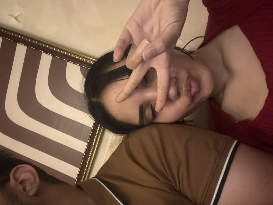 A woman with dark hair rests her head on a man's shoulder, her hand obscuring her face in a peace sign gesture. The man wears a brown shirt with a white stripe on the sleeve, only his ear visible. The woman wears a red shirt. Behind them is a framed wall decoration featuring brown and white stripes. The wall above the decoration appears cream-colored.