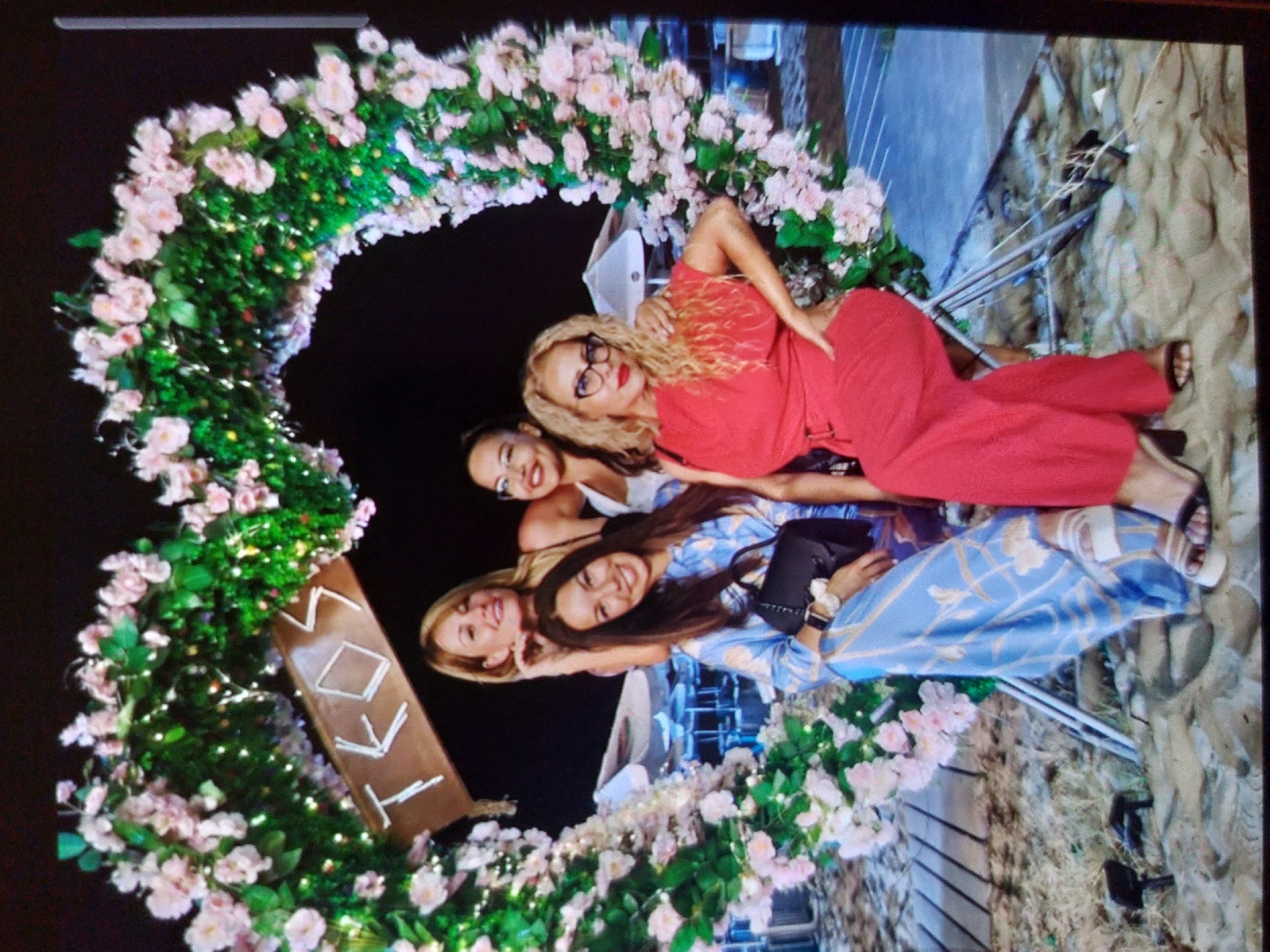 The image, taken in Tsarevo, Bulgaria, shows four women posing in an outdoor setting at night. The central feature is a large, heart-shaped archway covered in green foliage and numerous pink flowers, interspersed with delicate white string lights that illuminate the scene.
The four women are positioned within the arch, appearing happy and smiling at the camera. The woman at the top has blonde, curly hair, wears glasses, and is dressed in a bright red top and matching pants, reclining casually. Below her, a woman with darker hair wears a white top. Next to her, another woman with dark hair is dressed in a blue patterned long-sleeved garment, holding a small black bag. The fourth woman, with blonde hair, is partially visible at the bottom of the group.
The background is dark, indicating it is nighttime, and reveals hints of stone or rock walls and some metal structures, possibly railings or part of an outdoor building structure. A wooden sign is visible within the lower left curve of the heart arch, displaying the letters "IKKI". The overall mood is joyful and celebratory, suggesting a festive social gathering or event.