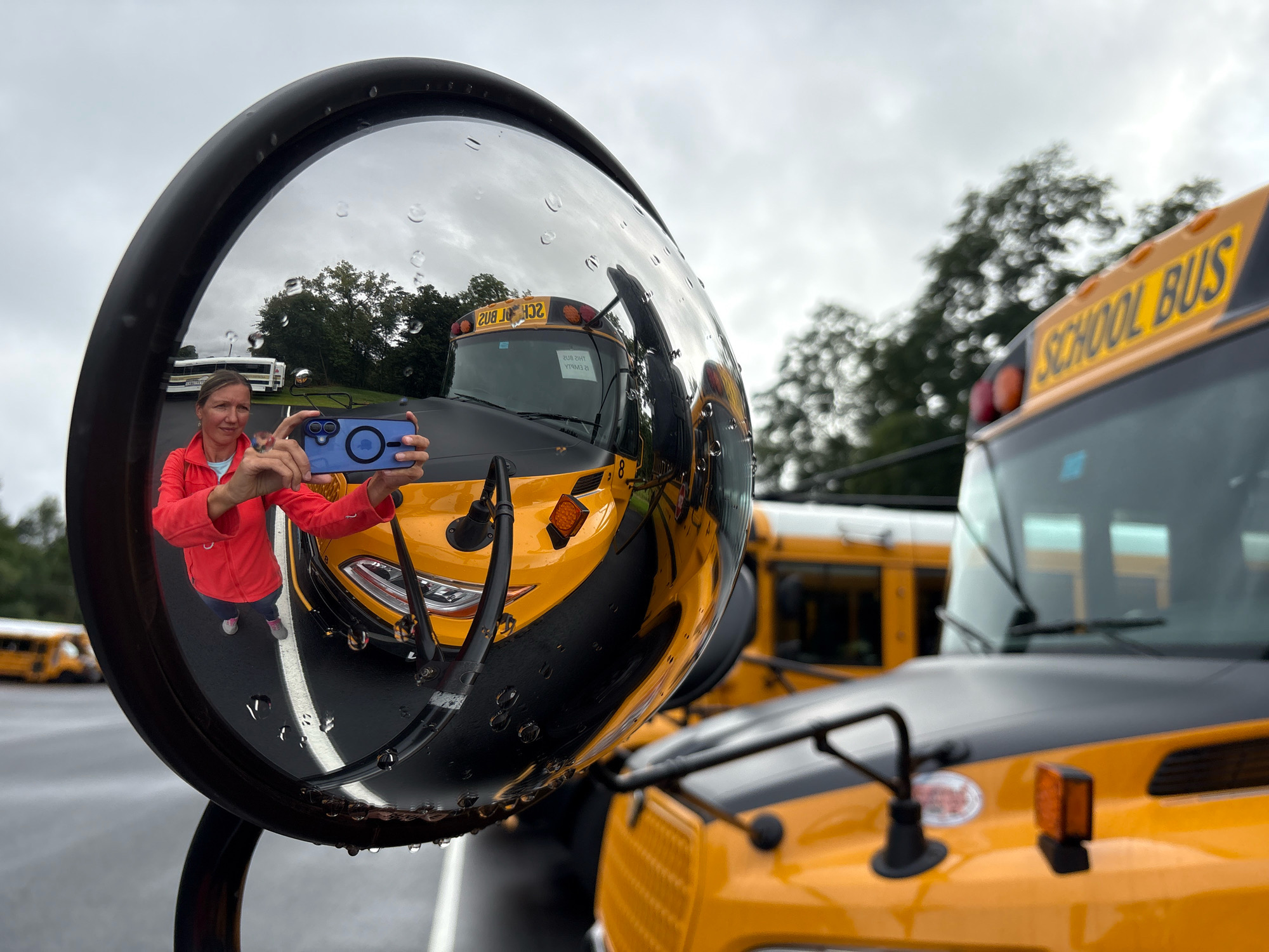 A woman in a red jacket and blue jeans is taking a selfie in the convex mirror of a school bus. She is holding a blue smartphone up to the mirror, and her reflection shows her taking the picture. The school bus is yellow, and the mirror is attached to the side of the bus. In the reflection, other school buses are visible, as well as trees and a cloudy sky. The image appears to have been taken outdoors in Newington, United States of America, as evidenced by the context of the school buses. There are raindrops on the mirror, suggesting it may be raining or has recently rained.
