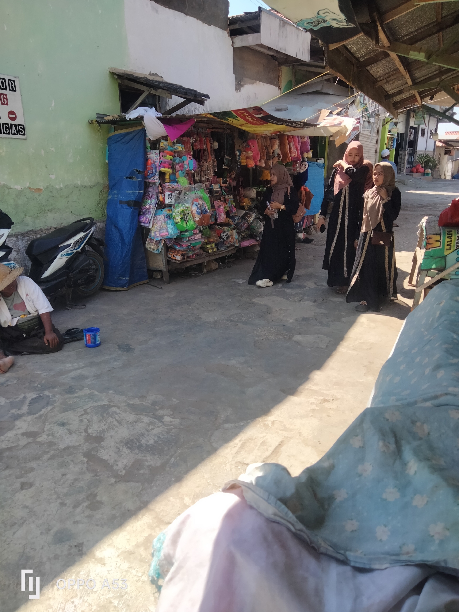 In Badung, Indonesia, a street scene unfolds with a small roadside stall selling an array of colorful toys and trinkets. Three women, dressed in traditional Islamic attire, are browsing the merchandise. One woman in the foreground, wearing a black abaya with gold trim, examines items on display. Behind her, two other women, also in dark abayas and hijabs, stand together, one holding a phone, perhaps taking a picture or communicating.

To the left of the stall, a person wearing a straw hat and a white t-shirt is seated on the ground, leaning against a parked motorcycle. A blue plastic cup sits near them, suggesting they might be collecting donations. The stall itself is a simple wooden structure covered by a corrugated metal roof and a blue tarp. Merchandise spills out onto the wooden shelves, creating a vibrant display.

The ground is a paved, grey concrete surface, showing signs of wear and tear. Sunlight casts distinct shadows across the scene, indicating it is likely daytime. In the background, other structures and a narrow street are visible, hinting at a densely populated urban area. The overall impression is one of a busy marketplace, with daily commerce and social interaction taking place under the Indonesian sun.