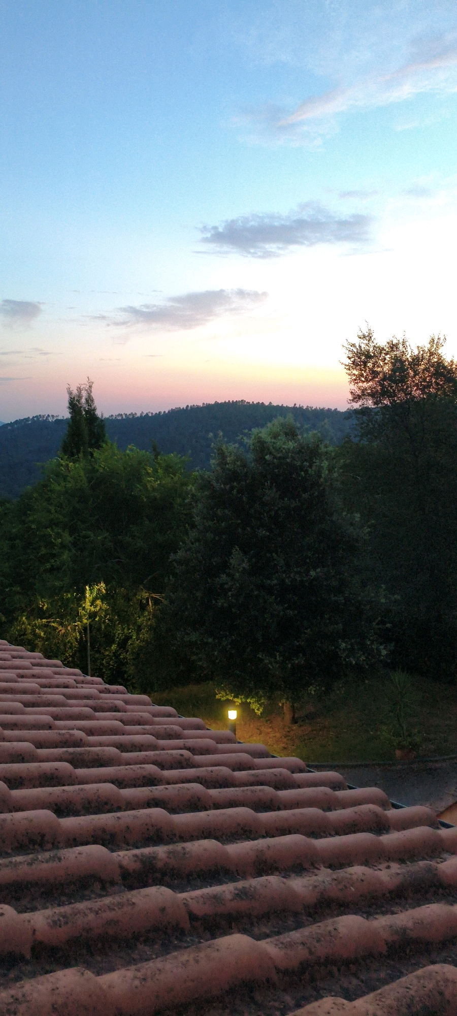 The image captures a twilight view over a tiled roof in Casale di Pari, Italy. The foreground is dominated by the texture of reddish-brown, weathered roof tiles, arranged in parallel rows. Beyond the roof, a dense line of dark green trees forms a natural barrier, separating the foreground from the distant landscape. This treeline follows the undulating contour of a hill.
The sky above transitions from a pale blue near the top to softer hues of pink and orange near the horizon, indicating the final moments of daylight. Scattered, wispy clouds are visible against the lighter sky. Two small, illuminated garden lights are present in the mid-ground, casting a warm, yellow glow on the surrounding foliage and the ground. One light is positioned among the trees to the left, and another is closer to the right edge of the frame, near the base of a tree. The overall impression is one of serene natural beauty transitioning into evening.