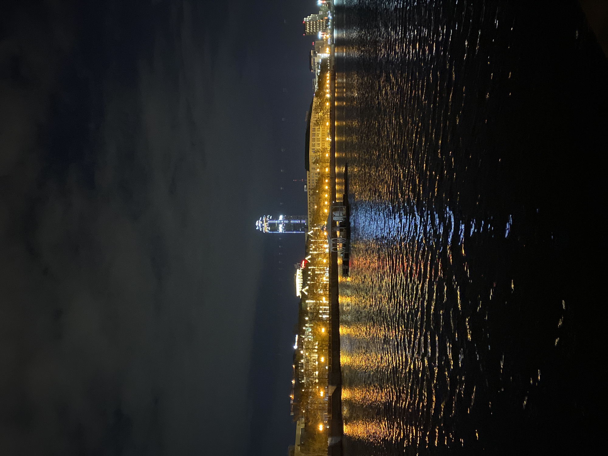 The image shows a nighttime view of the Iset River in Yekaterinburg, Russia. The cityscape is reflected in the water, with visible buildings, streetlights, and the prominent Vysotsky skyscraper. The sky is dark with some cloud cover. The overall scene is dimly lit by city lights.