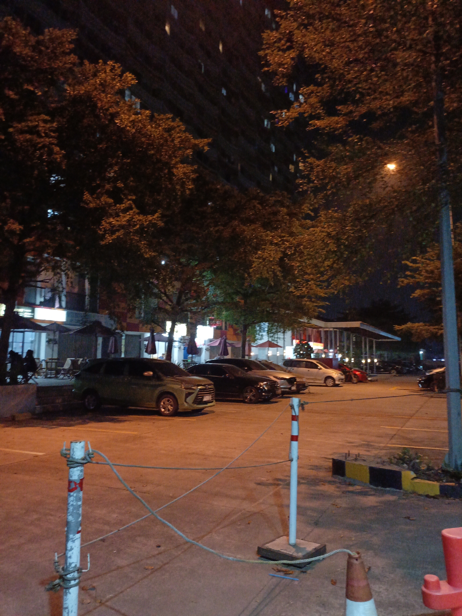 The image shows a dimly lit outdoor parking lot at night. Several cars are parked in the lot, including a light green SUV in the foreground. Trees with autumnal foliage cast shadows across the asphalt. In the background, there is a tall building with illuminated windows and a low-rise structure with awnings and outdoor seating areas, possibly a restaurant or cafe. The lighting in the scene comes from streetlights and internal building lights, creating a warm, orange glow. A white and red striped pole with ropes attached is visible in the foreground, suggesting a boundary or a temporary closure. The overall environment appears to be an urban or semi-urban setting in Wangunharja, Indonesia, likely during the evening hours.
