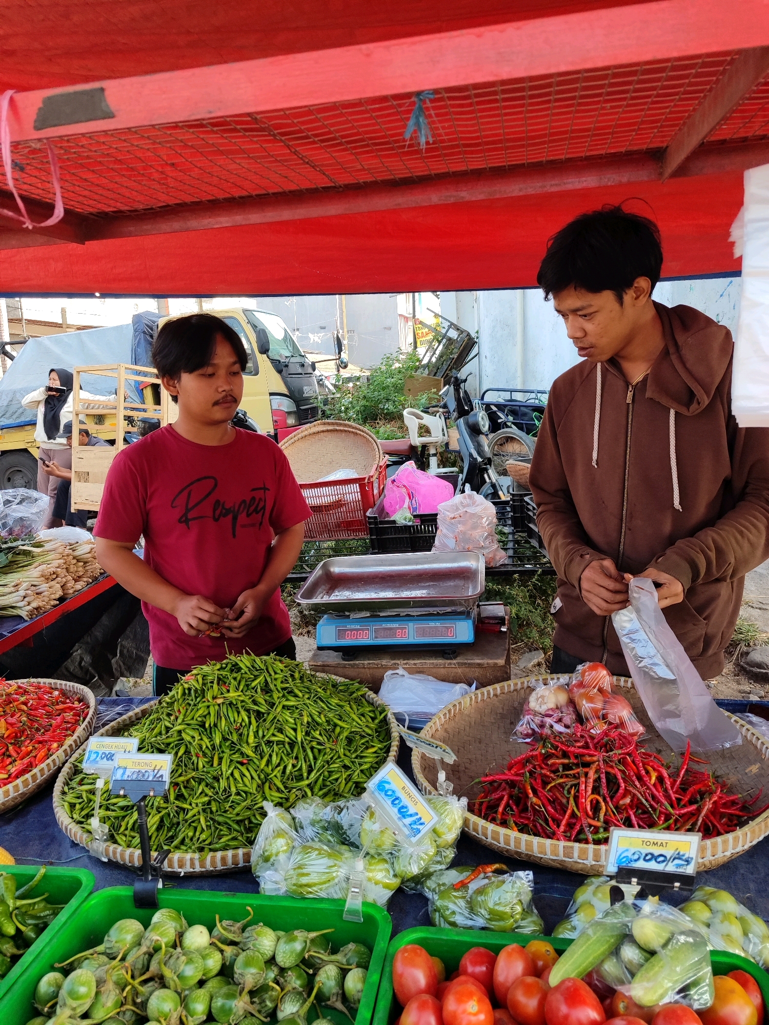 Two young men are standing behind a market stall laden with produce. The man on the left, wearing a red t-shirt with "Respect" printed on it, is looking down and holding some small red chilies in his hands. The man on the right, in a brown hooded jacket, is holding a clear plastic bag and appears to be preparing to bag some items.
In front of the men are several woven baskets and green plastic crates filled with various vegetables. One large woven basket is overflowing with small green chilies. Another basket contains red chilies and some onions and tomatoes. In the foreground, green plastic crates hold items that appear to be eggplants and tomatoes. There are also bags of produce, some with price tags visible, indicating items like "Gengek Hijau" (green chilies) and "Tomat" (tomatoes) with prices. A blue digital scale is also on the stall.
Behind the stall, a yellow truck with a blue tarp is visible, along with other market activity. The stall is covered by a red awning. The overall scene depicts a busy outdoor market environment. The presence of specific Indonesian produce and the visual context strongly suggest this location is a traditional market in the city of Garut, Indonesia.